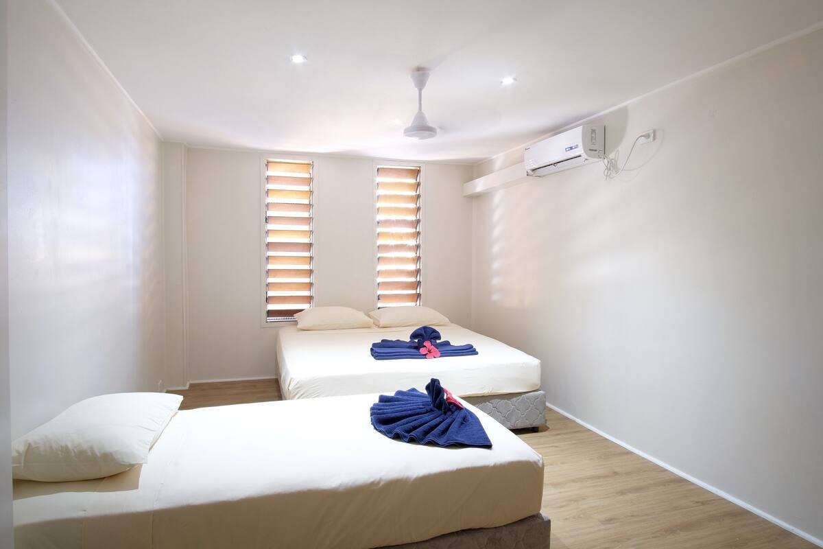 A hotel room with two single beds, a ceiling fan, an air conditioning unit, and a window with blinds.