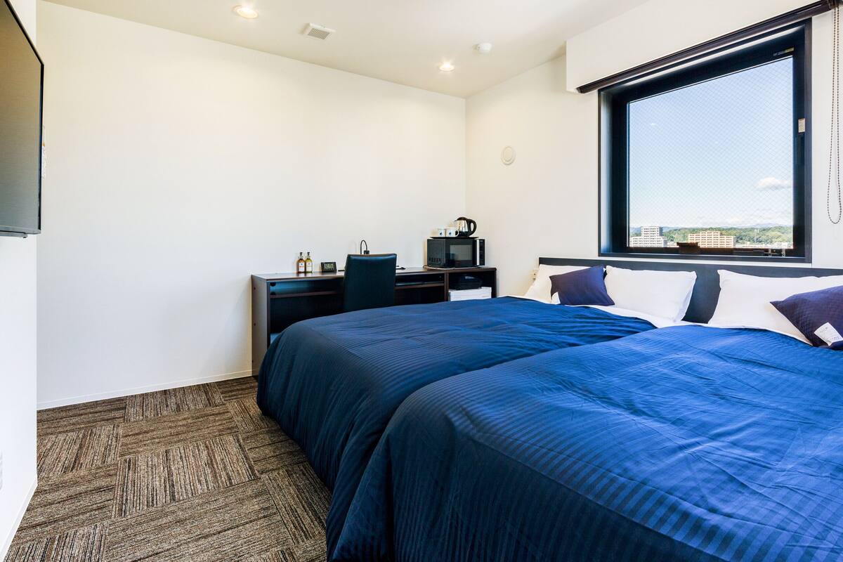 A hotel room with a large bed, a desk with a computer and a kettle, a window with a city view, and a carpeted floor.