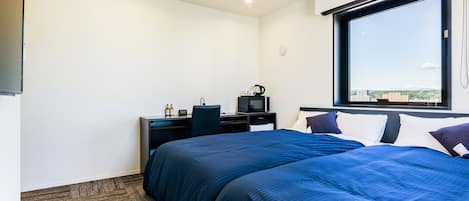 Una habitación de hotel con una cama grande, un escritorio con computadora y hervidor, una ventana con vista a la ciudad y piso alfombrado.