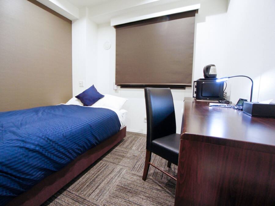 A hotel room with a single bed, a desk with a computer, and a window with a blind.