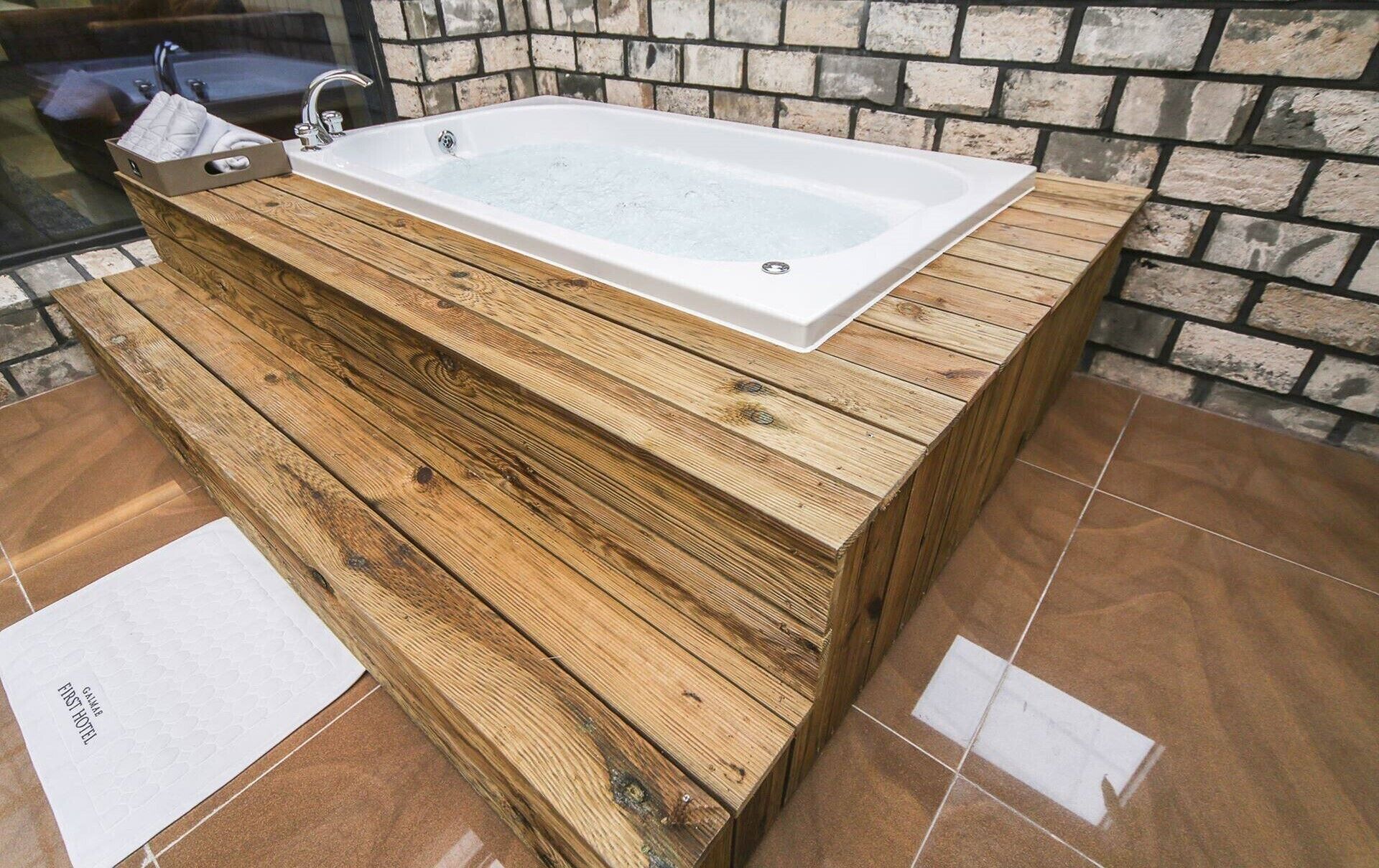 Una bañera rectangular con base de madera, frente a una pared de piedra.