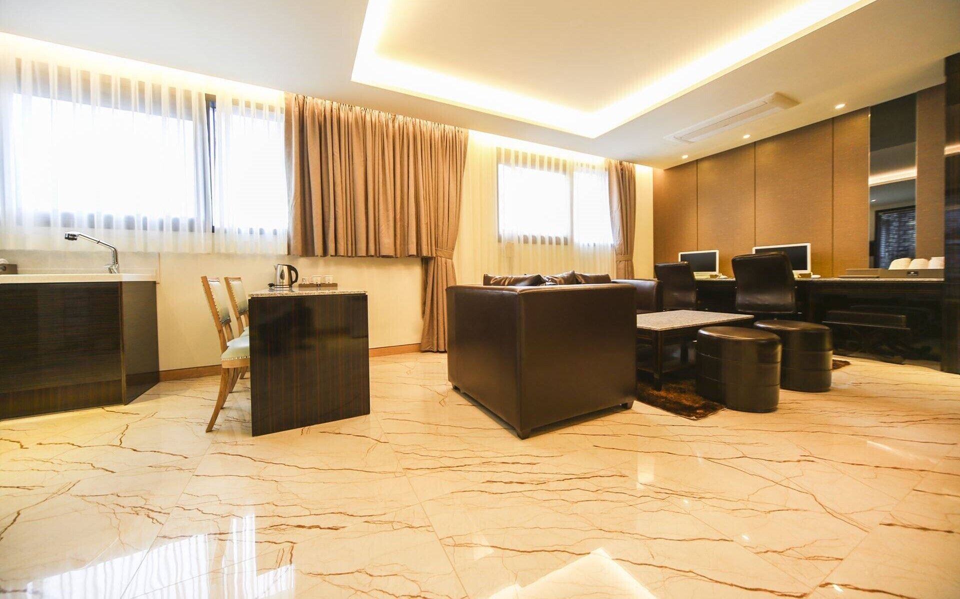 A modern hotel room with a marble floor, a seating area with a sofa and chairs, a small table with a kettle, and a desk with computers.