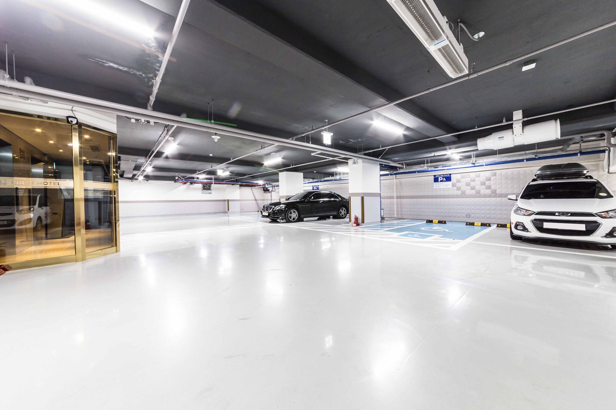 A clean, well-lit parking garage with a glass entrance, a white car, and a black car parked inside.