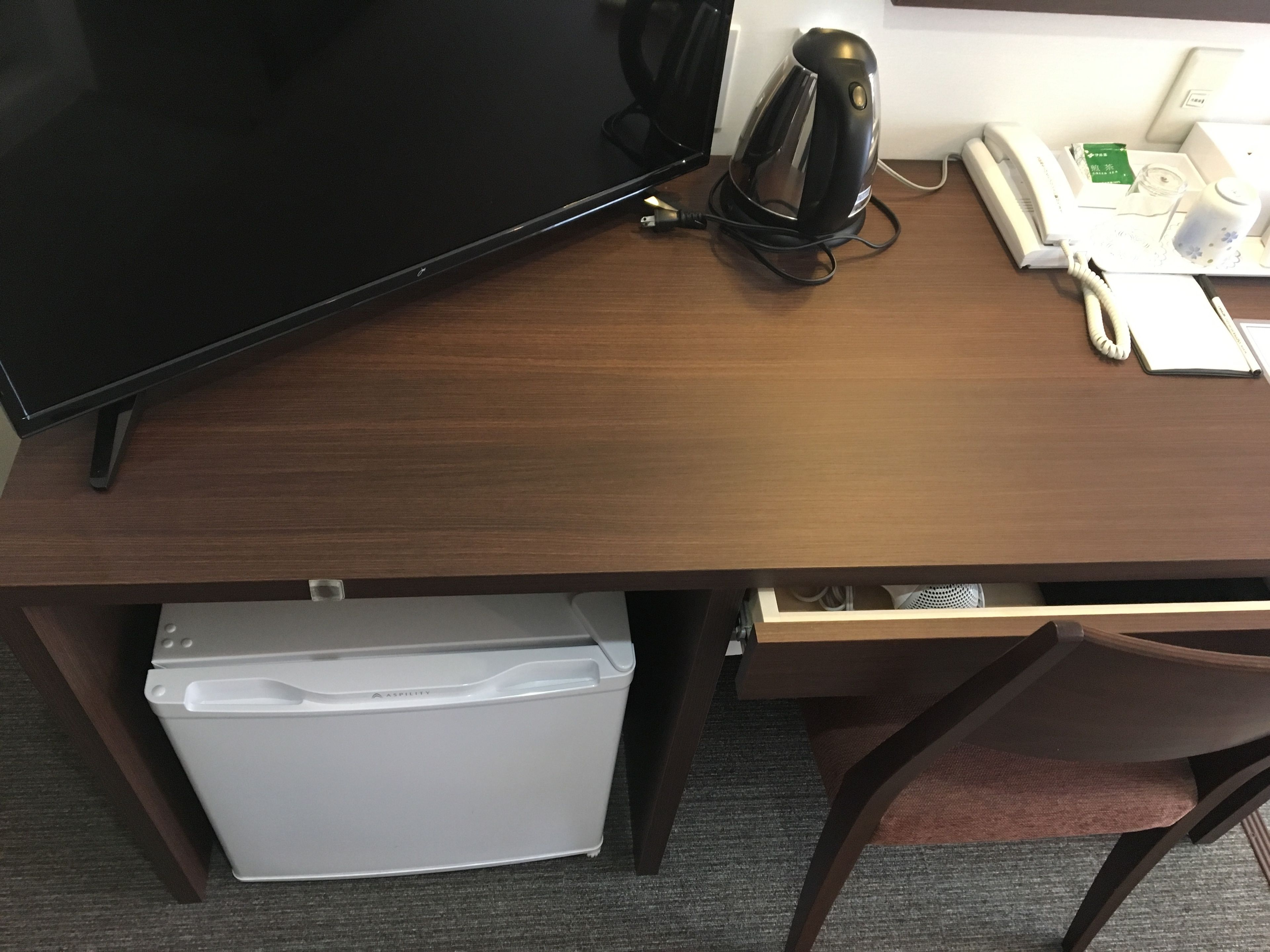 Un escritorio de madera con un televisor de pantalla plana, una tetera eléctrica negra, un teléfono blanco y un pequeño electrodoméstico blanco.