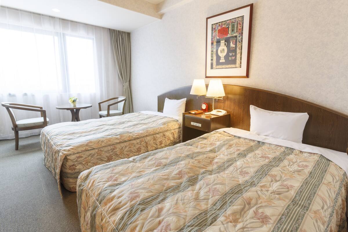 Ein Hotelzimmer mit zwei Betten, einem kleinen Tisch, einem Stuhl, einem Nachttisch mit Lampe und einem gerahmten Bild an der Wand.