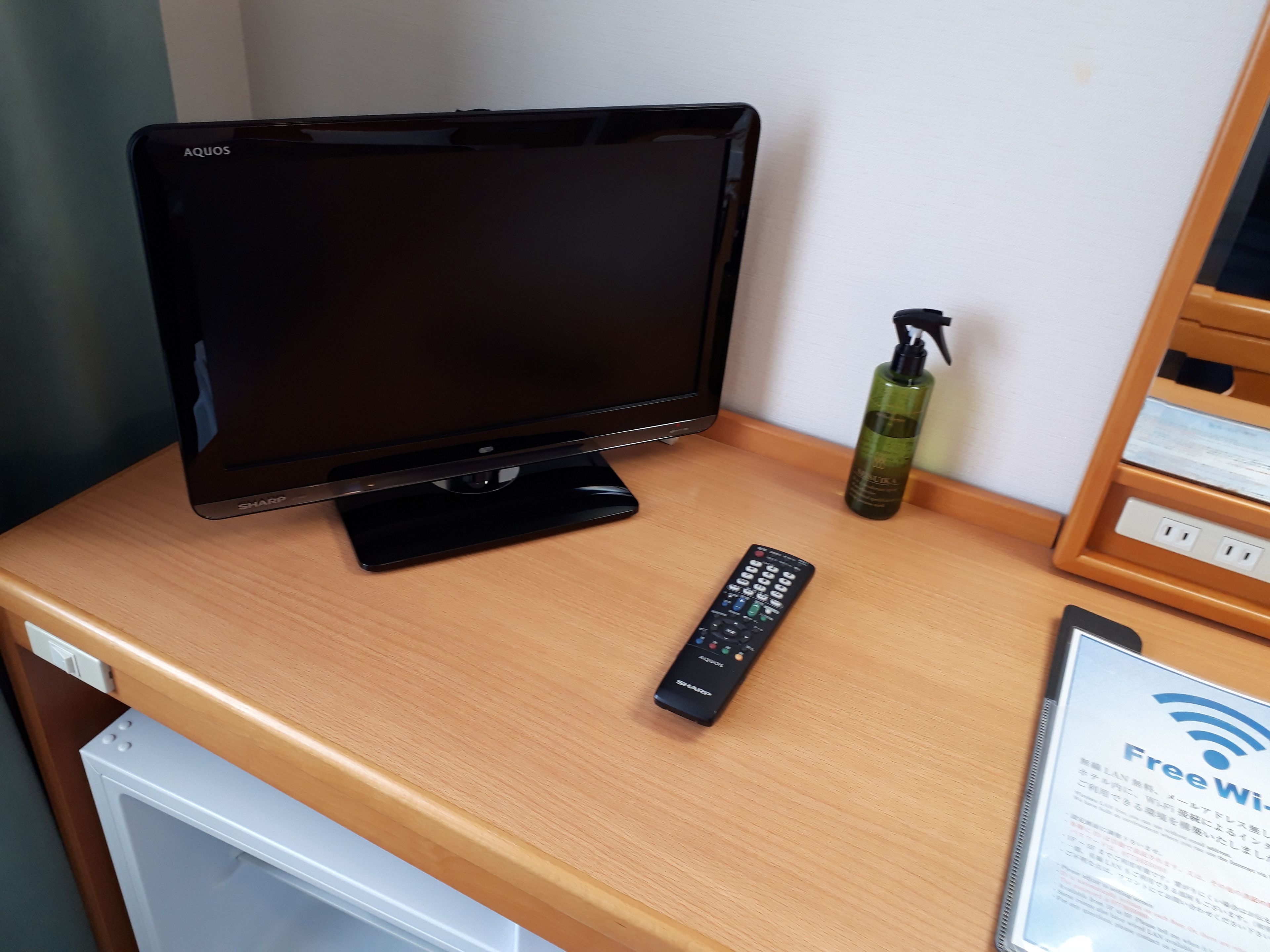 A hotel room with a flat-screen TV, a remote control, a bottle of hand sanitizer, and a sign indicating free Wi-Fi.