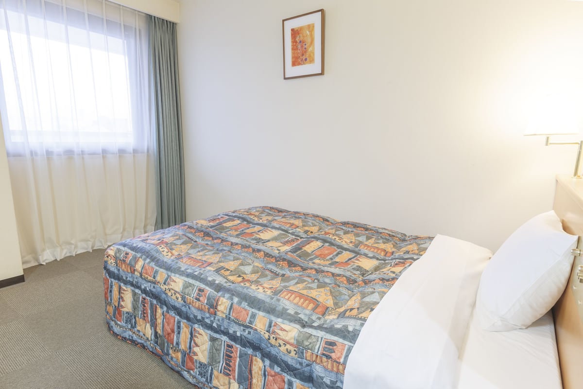 Ein Hotelzimmer mit Bett, Fenster mit Vorhängen, einem gerahmten Bild an der Wand und einer Nachttischlampe.