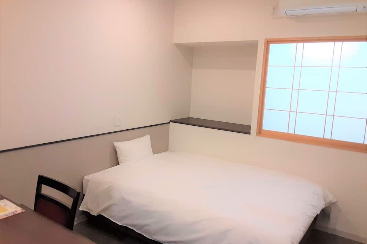 Ein kleines, sauberes Zimmer mit einem Einzelbett, einem Sessel, einem Tisch und einem Fenster mit Jalousien.