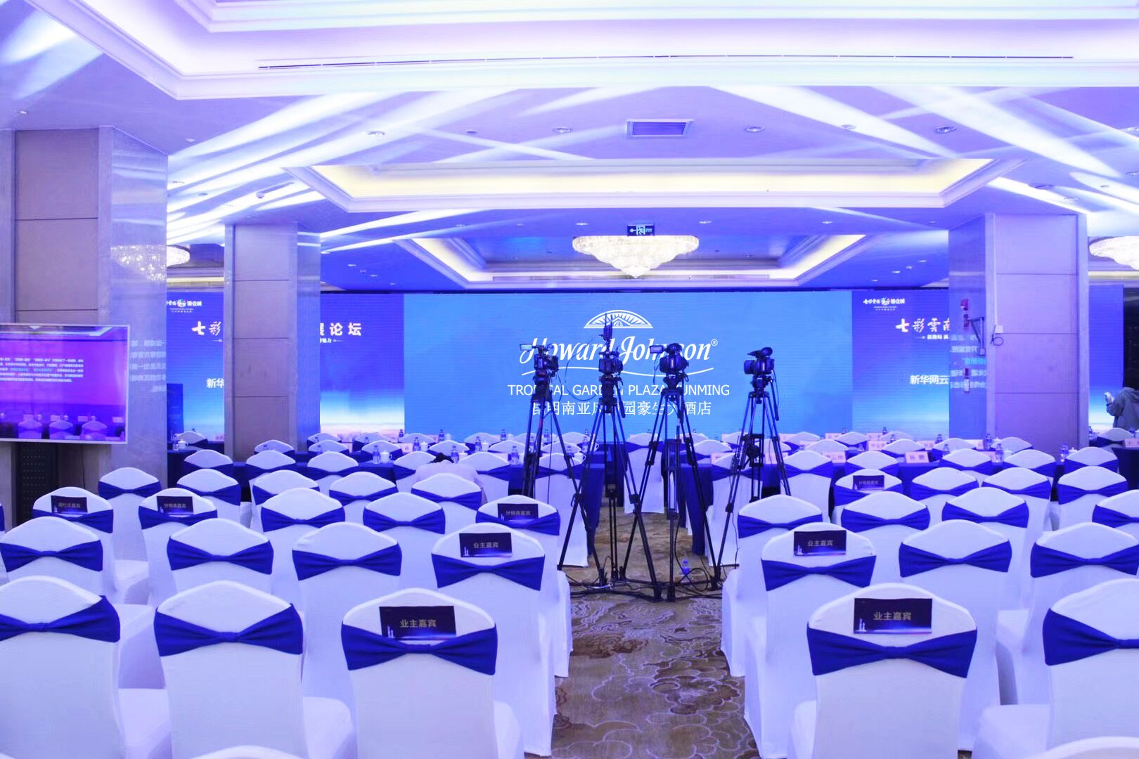Un salón de conferencias con filas de sillas blancas con cintas azules, un escenario con fondo azul y el logotipo de 'Tropical Garden Plaza'.