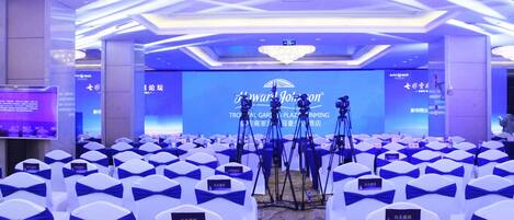 Un salón de conferencias con filas de sillas blancas con cintas azules, un escenario con fondo azul y el logotipo de 'Tropical Garden Plaza'.