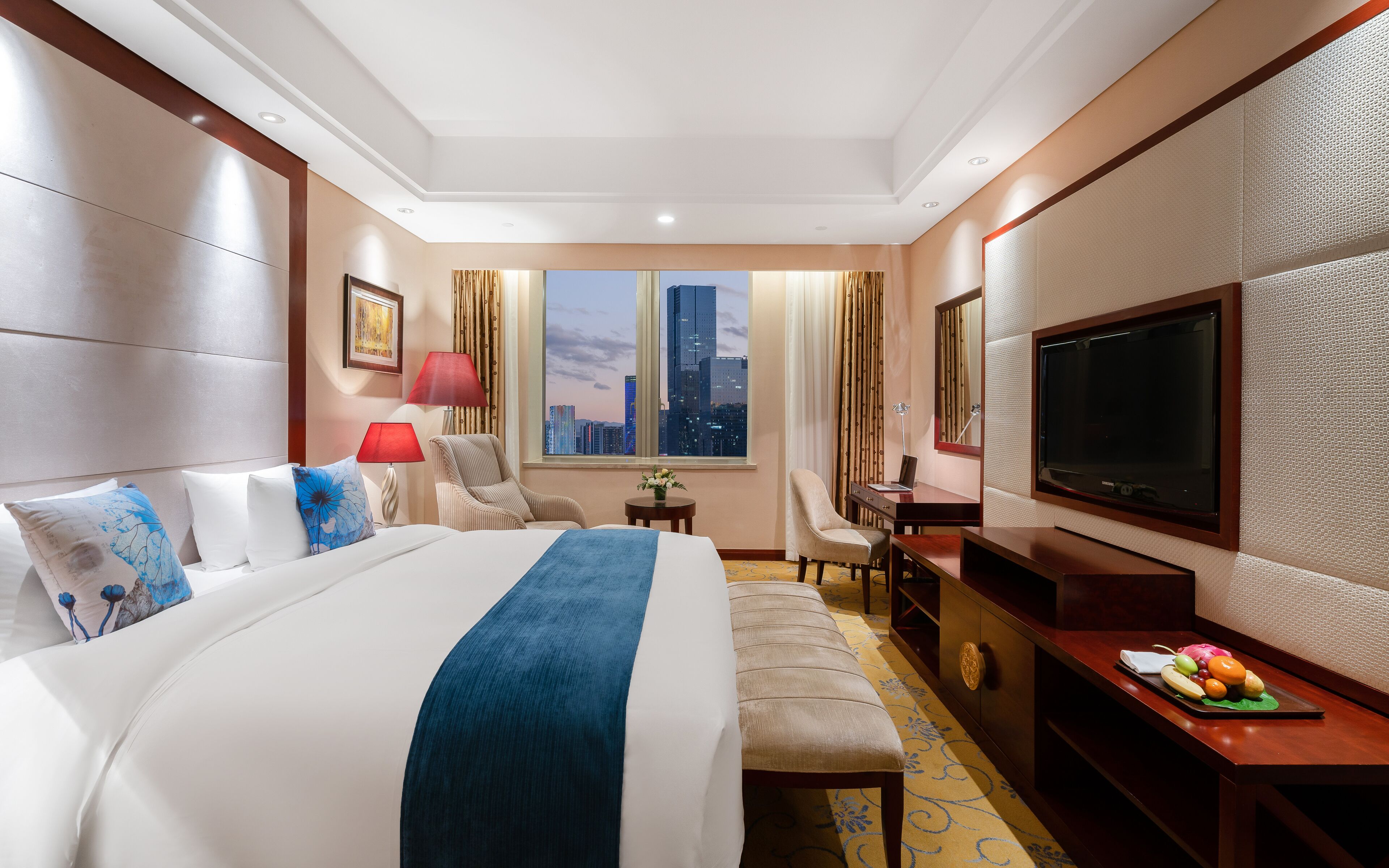 A hotel room with a large bed, a desk with a TV, a chair, and a view of the cityscape.