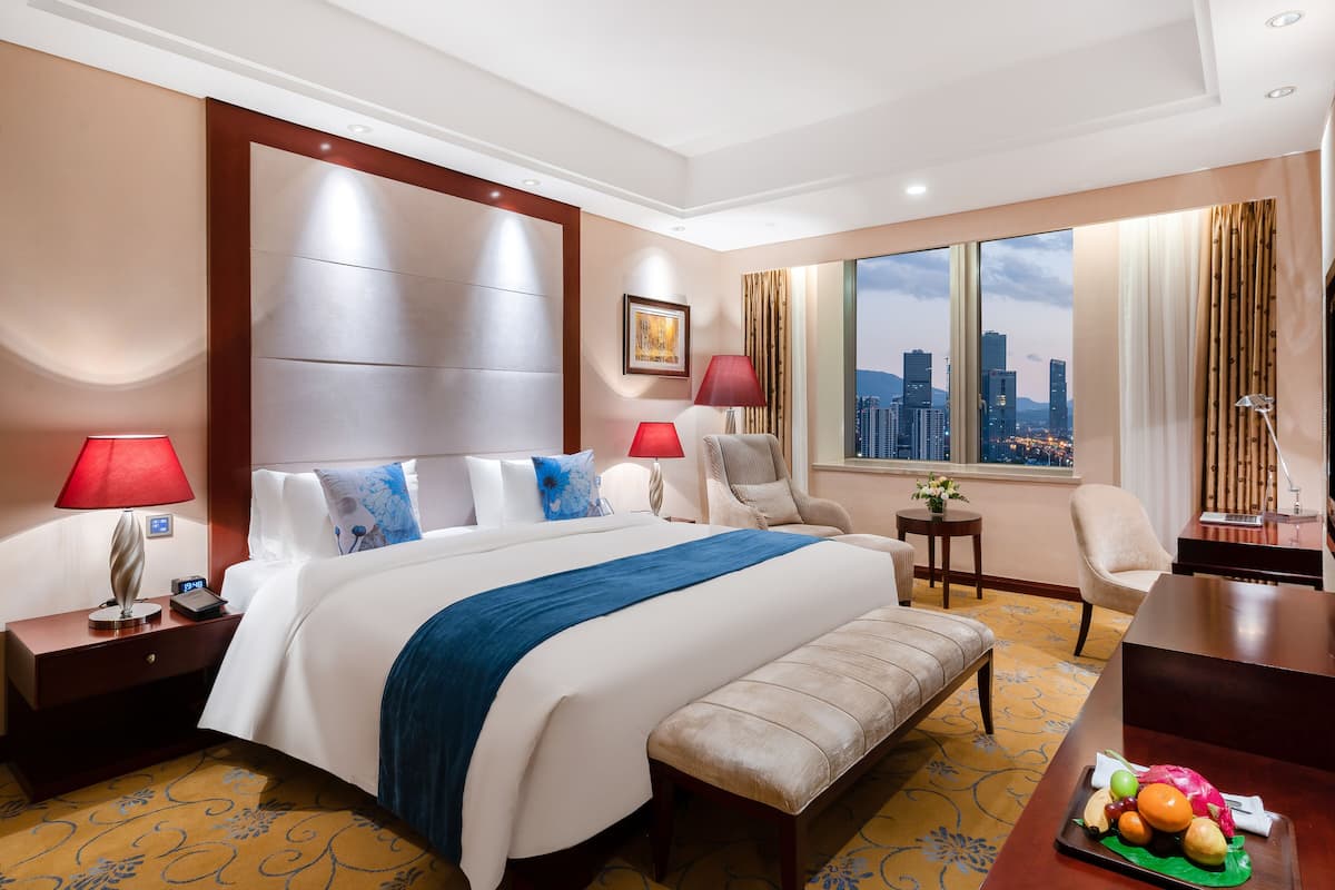 Ein Hotelzimmer mit einem großen Bett, zwei Nachttischen, einem Schreibtisch, einem Sessel, einer Bank, einem Fernseher und einem Fenster mit Blick auf die Stadt.