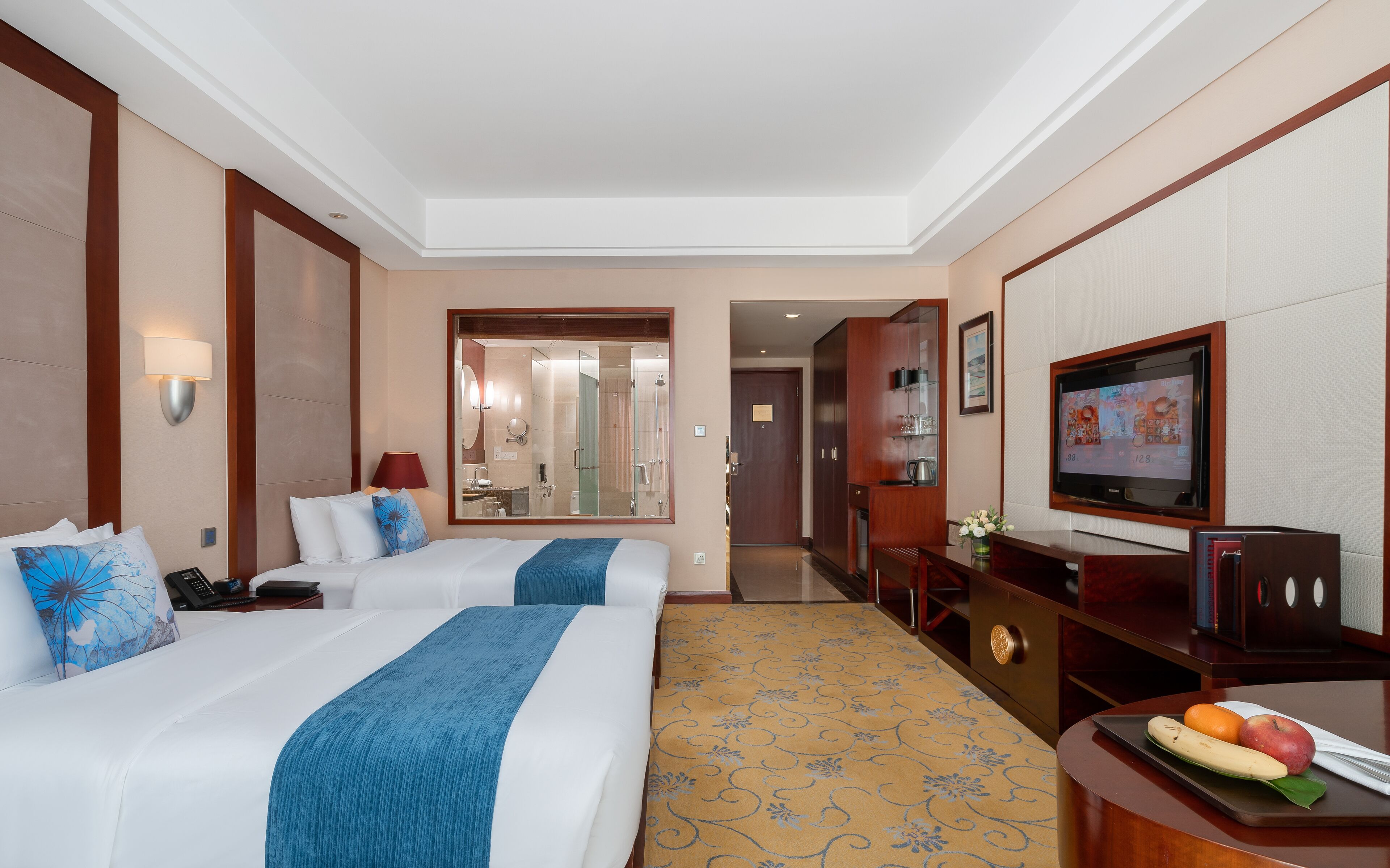 Habitación de hotel con dos camas, televisión, escritorio con frutero y baño visible a través de una puerta abierta.