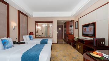 Habitación de hotel con dos camas, televisión, escritorio con frutero y baño visible a través de una puerta abierta.