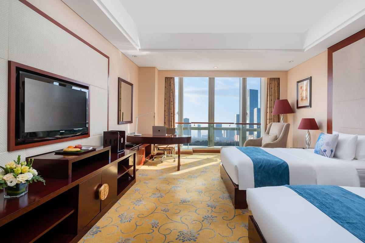 Ein Hotelzimmer mit großem Fernseher, einem Schreibtisch mit Laptop und einem Bett mit blauer Decke, mit Blick auf die Stadtlandschaft.