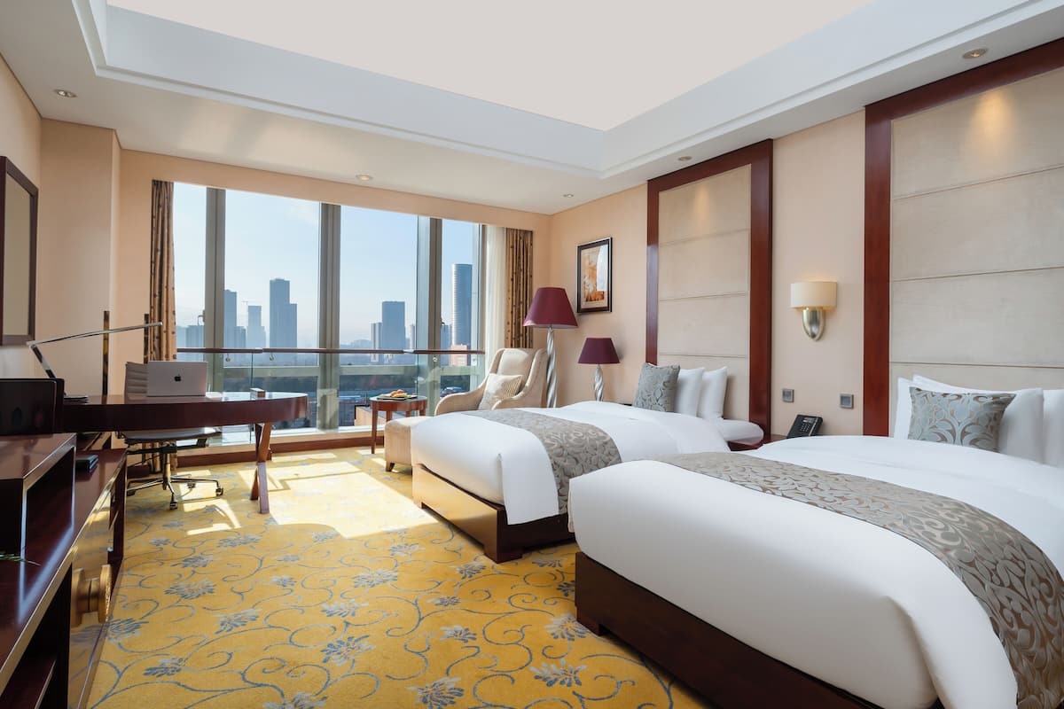 Ein Hotelzimmer mit zwei Betten, einem Schreibtisch, einem Stuhl, einer Lampe und Blick auf die Stadtlandschaft.