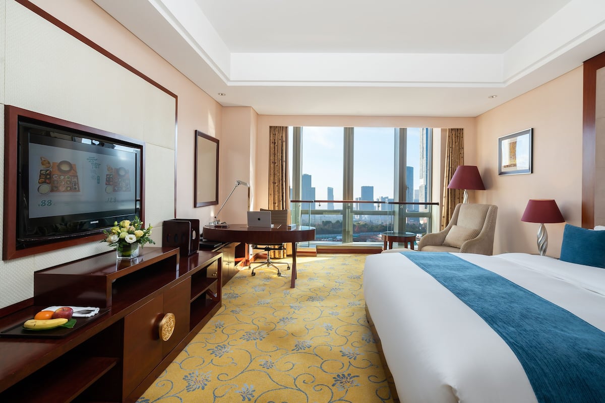Ein Hotelzimmer mit einem großen Bett, einem Schreibtisch mit einem Laptop, einem Fernseher und Blick auf die Stadtlandschaft.