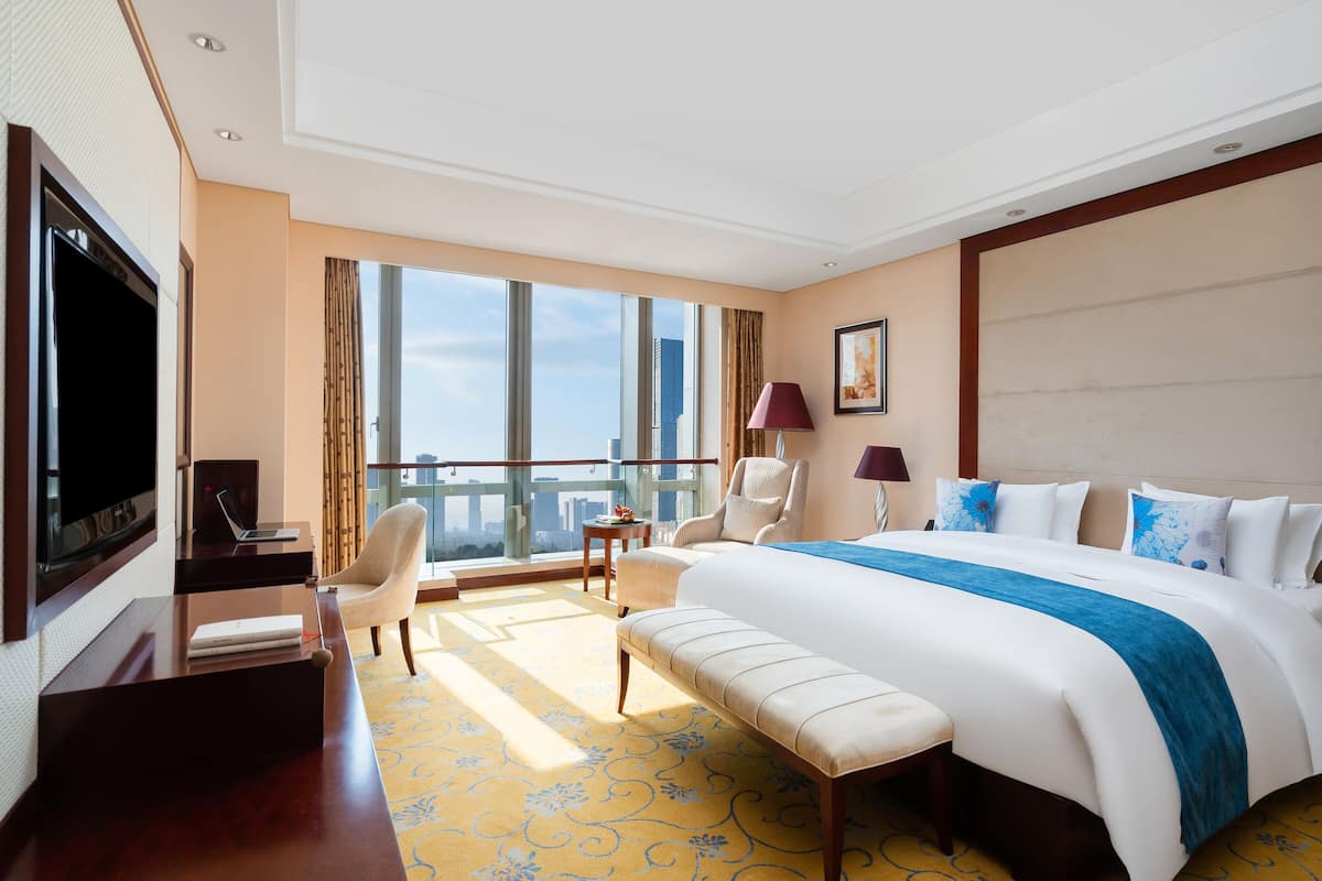 Ein Hotelzimmer mit einem großen Bett, einem Schreibtisch mit einem Laptop, einem Sessel, einem Fernseher und einem Balkon mit Blick auf die Stadt.