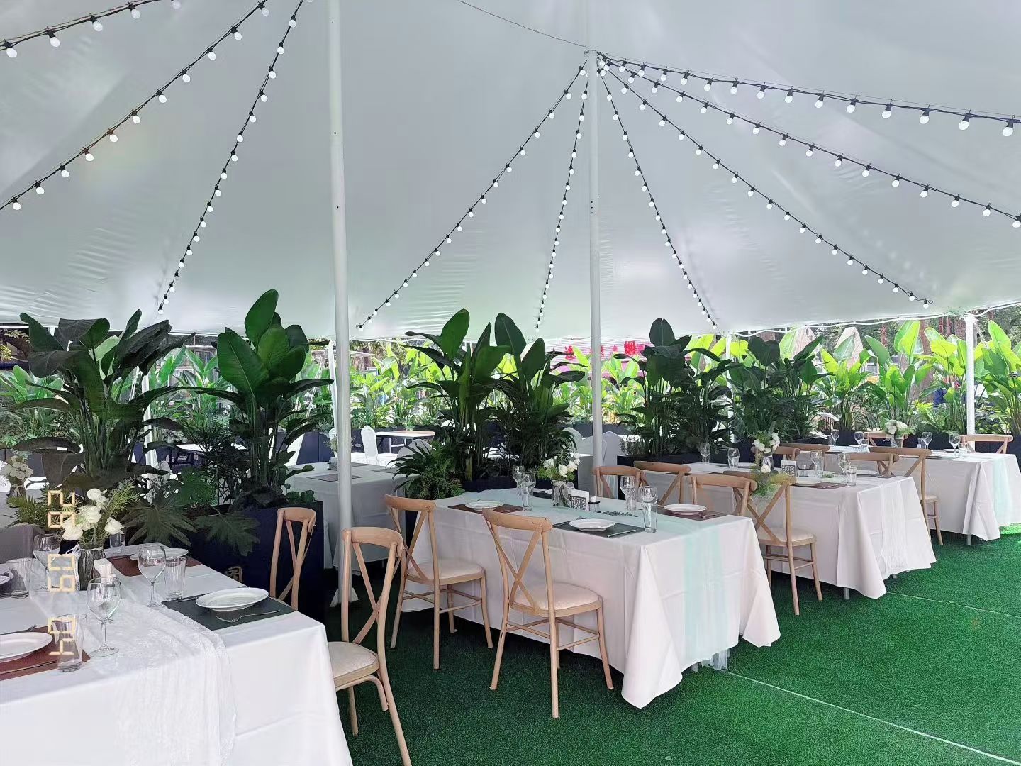 Un espacio al aire libre para eventos, con mesas dispuestas para comer, rodeado de vegetación e iluminación con luces colgantes.