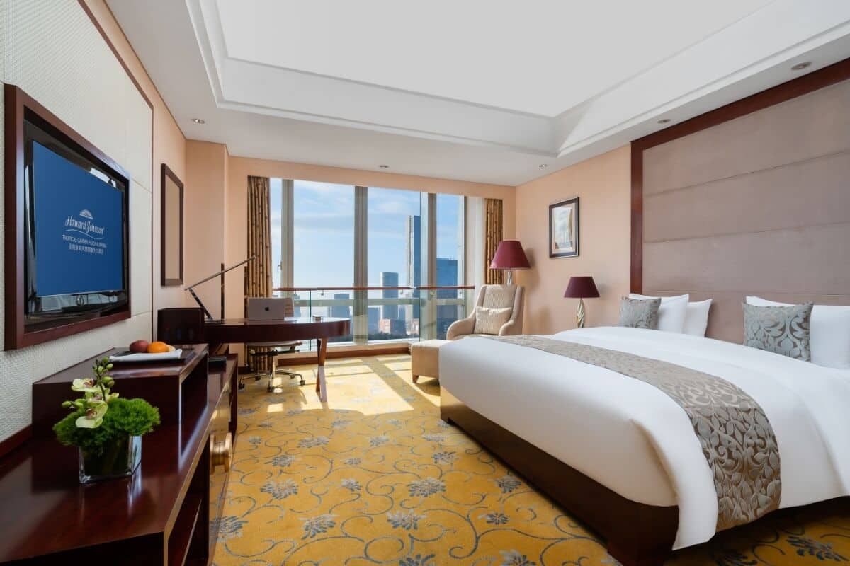 A hotel room with a large bed, a desk, a chair, a TV, and a view of the city.