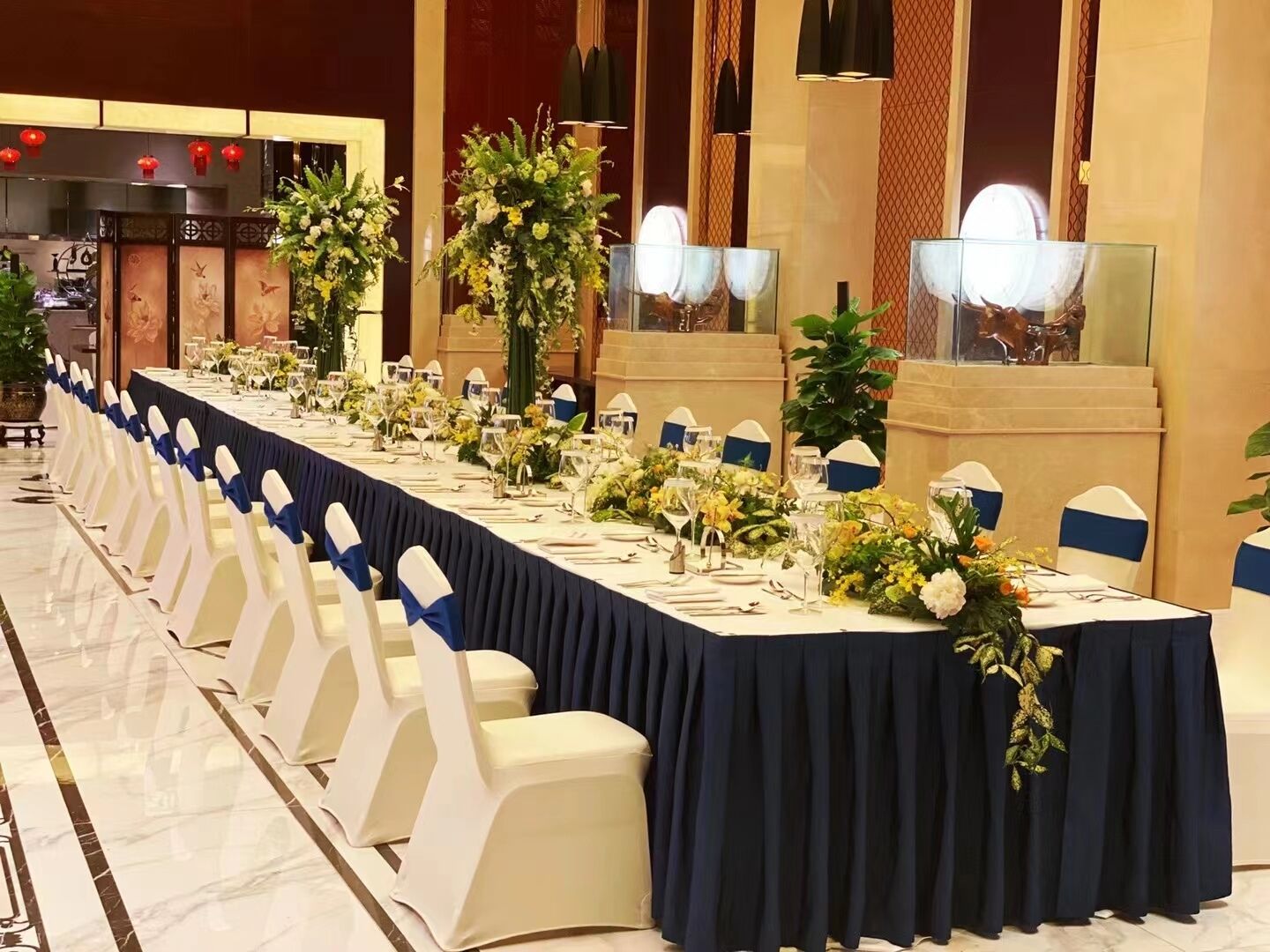 Un salón de banquetes con largas mesas dispuestas para un evento formal, decorado con centros de mesa florales y sillas blancas con cintas azules.