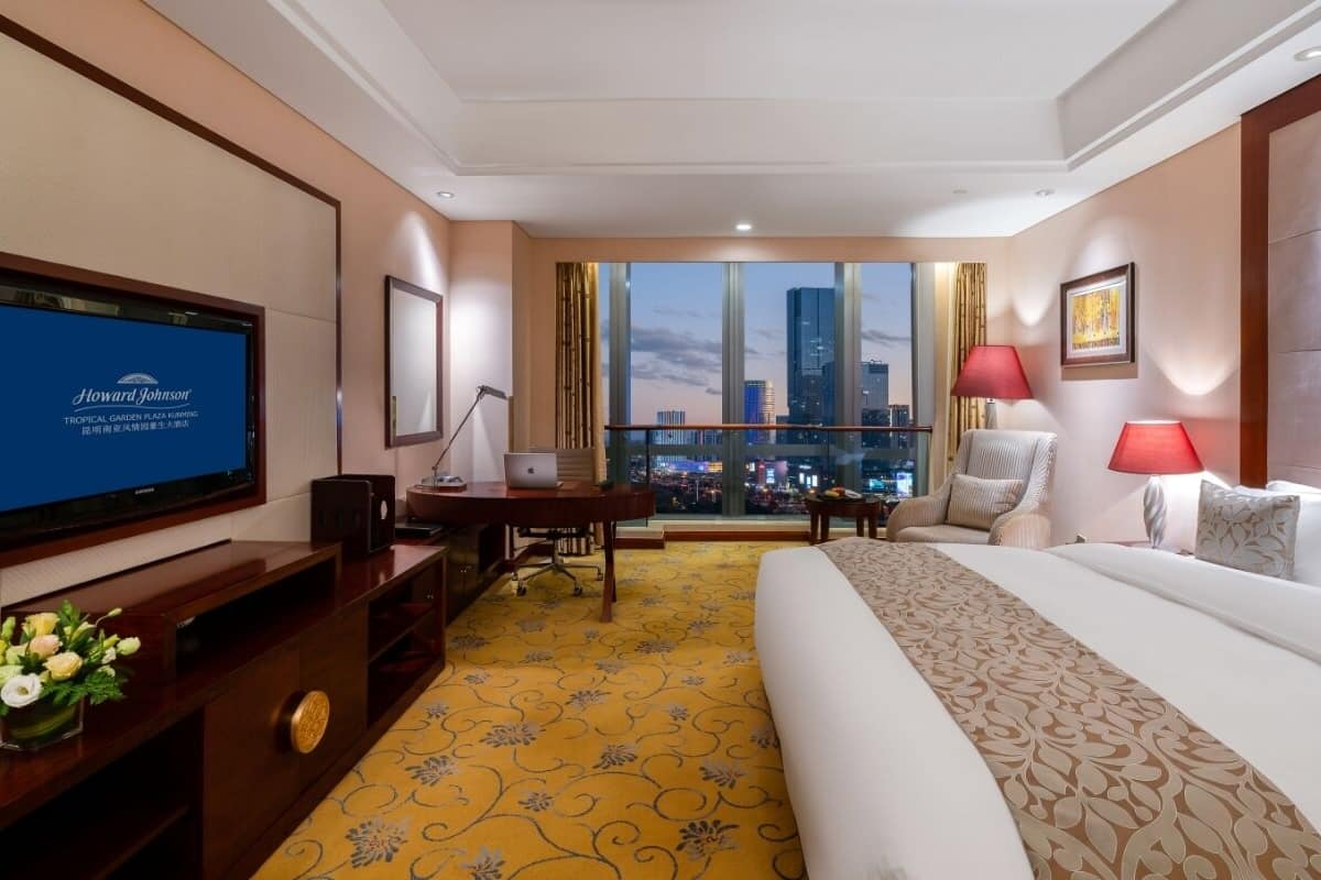 Ein Hotelzimmer mit einem großen Bett, einem Schreibtisch mit einem Laptop, einem Fernseher und Blick auf die Skyline der Stadt.
