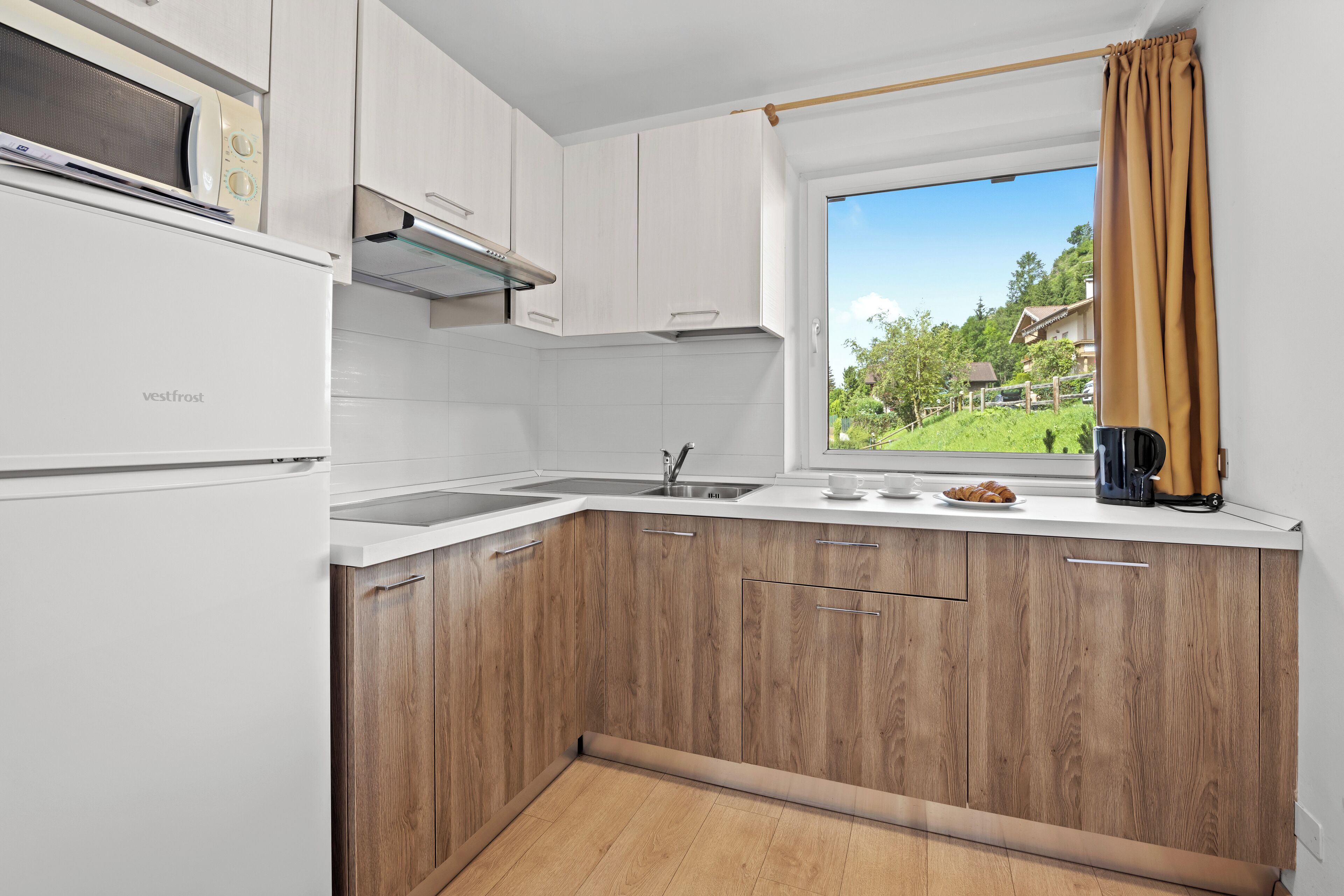 A modern kitchen with white cabinets, a wooden countertop, a built-in microwave, and a window with a view of a green landscape.