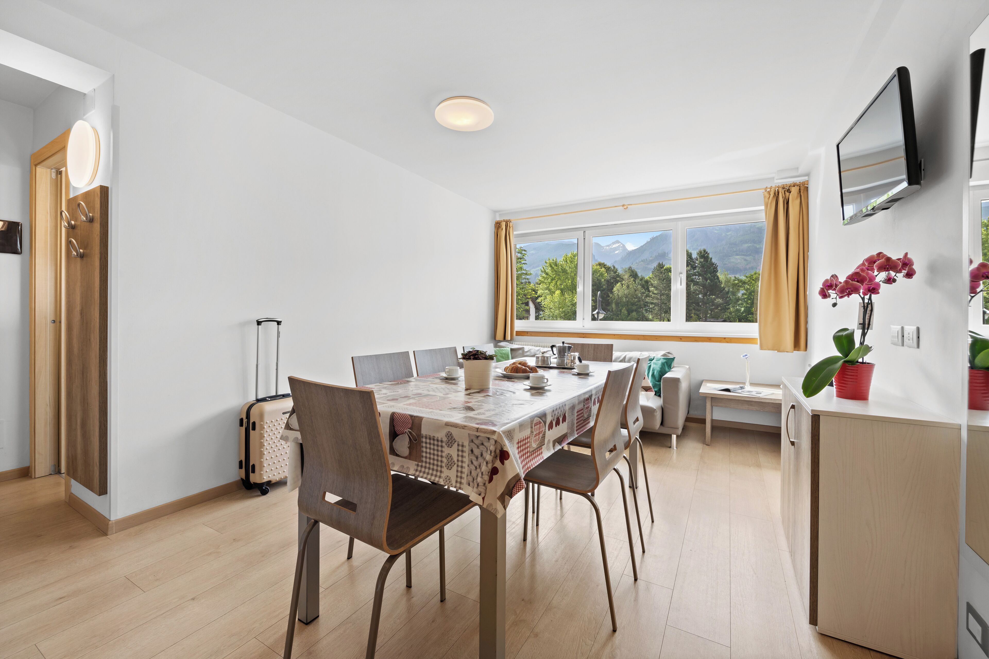 A modern dining area with a table set for four, a view of mountains through the window, and a TV mounted on the wall.