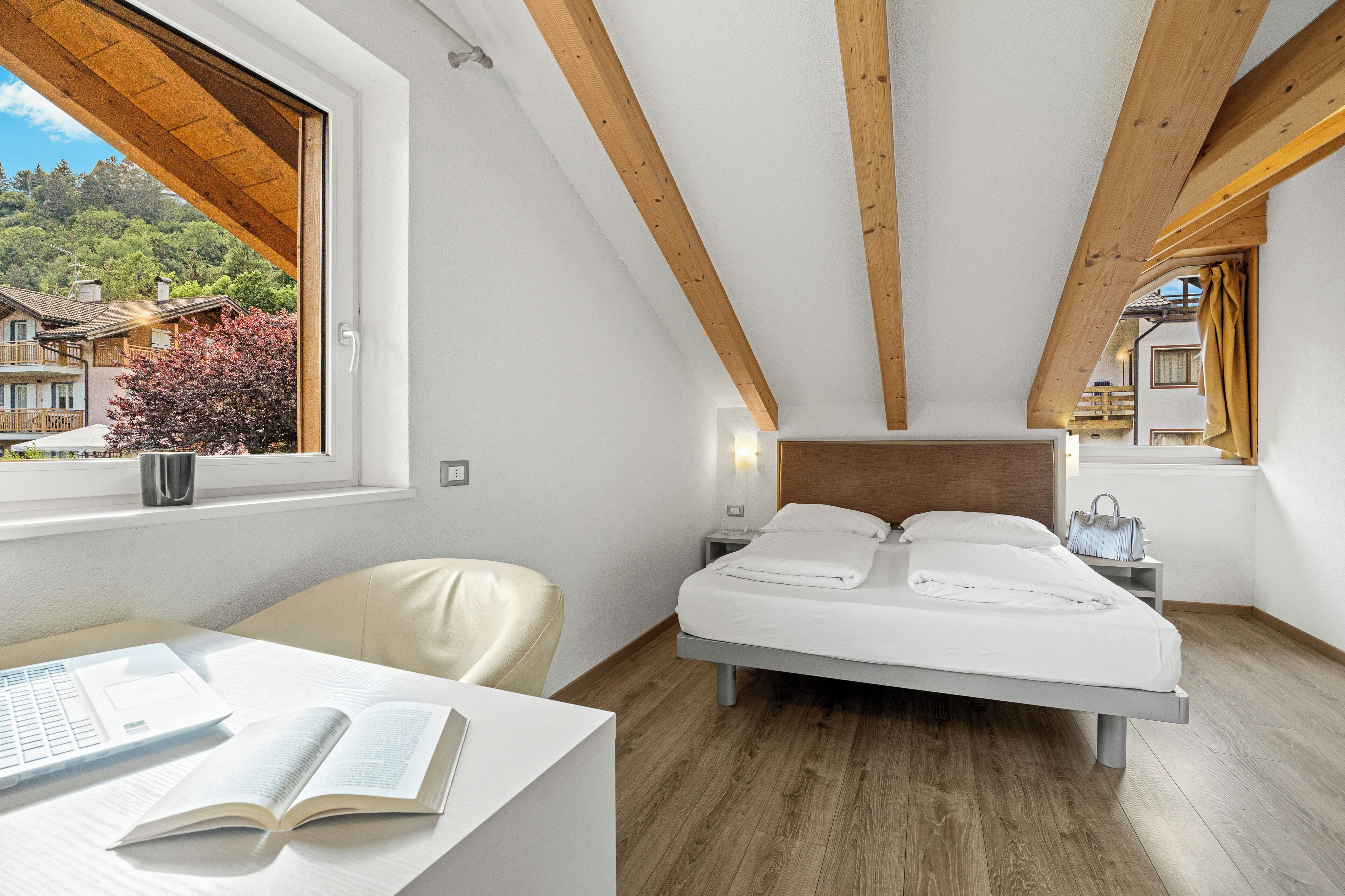 Un dormitorio con cama, escritorio con computadora portátil y libro, ventana con vista a casas y árboles, y vigas de madera en el techo.