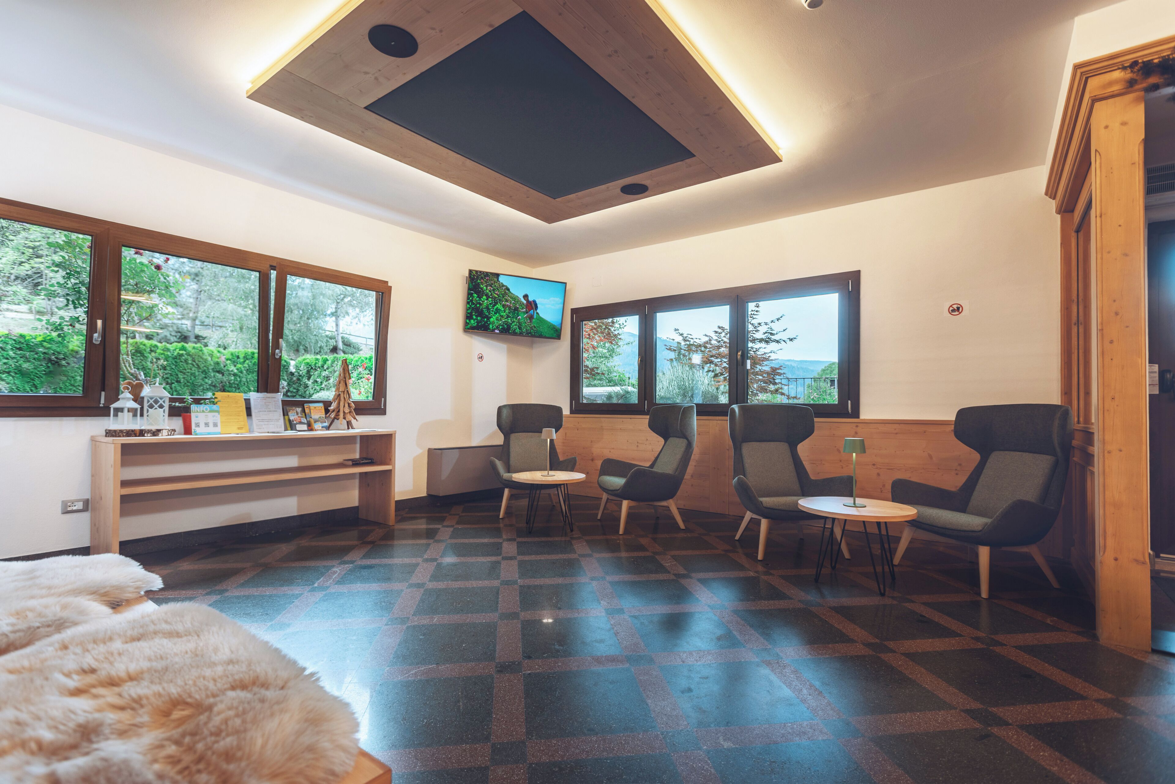 A modern room with a checkered floor, a wooden desk, two chairs, a television, and large windows with a view of greenery.