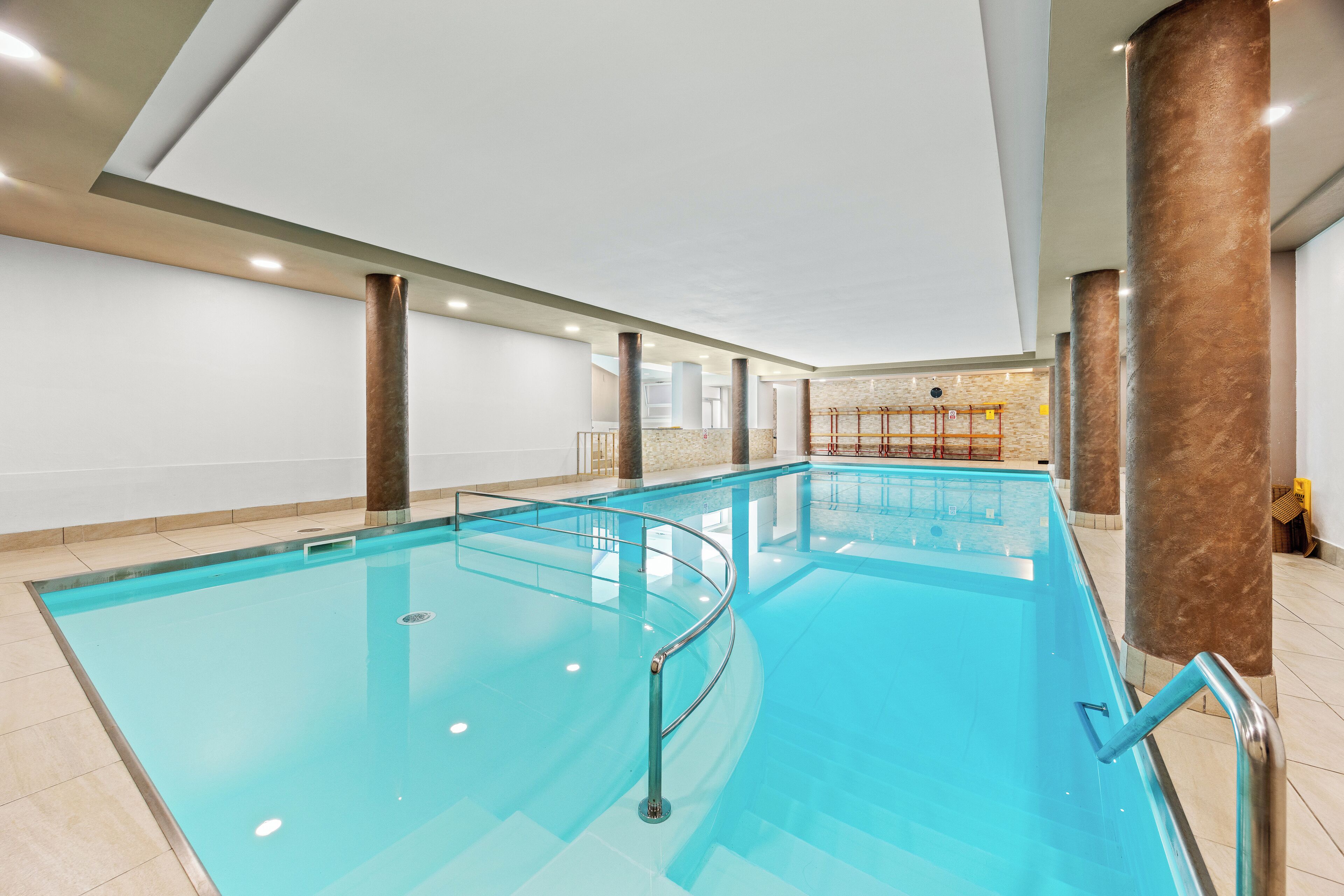 A large indoor swimming pool with clear water, surrounded by beige tiles and metal railings.