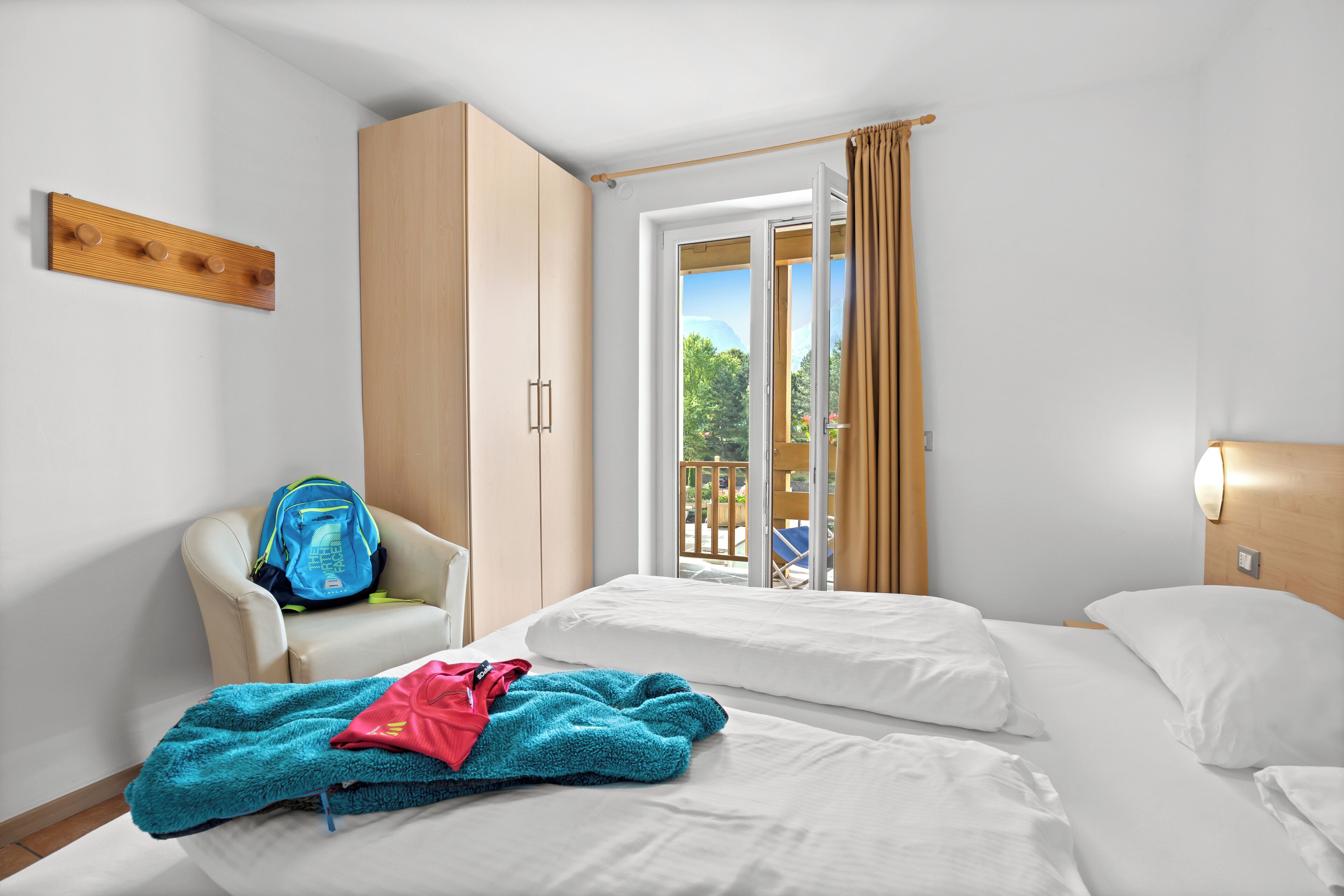 A hotel room with two beds, a chair, a wardrobe, and a view of the outdoors.