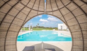 A pool with a clear view through a woven arch, surrounded by lounge chairs and a white building in the background. - Masseria Elysium (Mesagne)