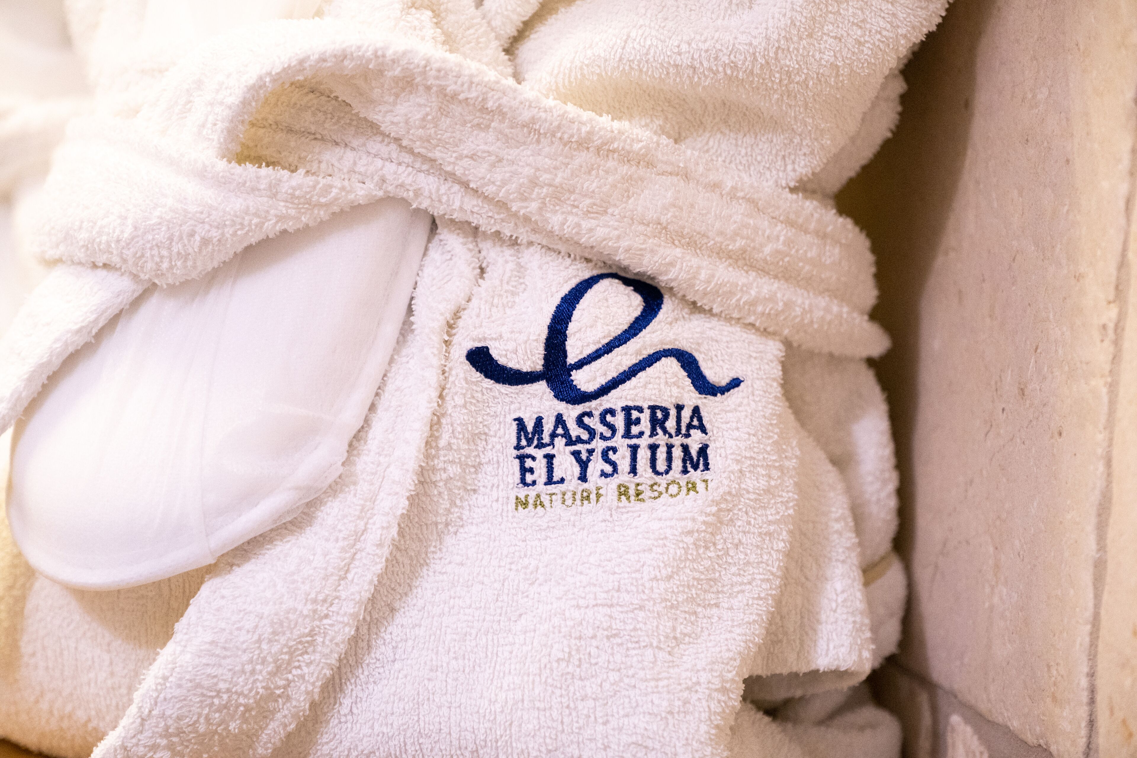 Una toalla blanca con el logo y el nombre "Masseria Elyium Nature Resort" bordados.