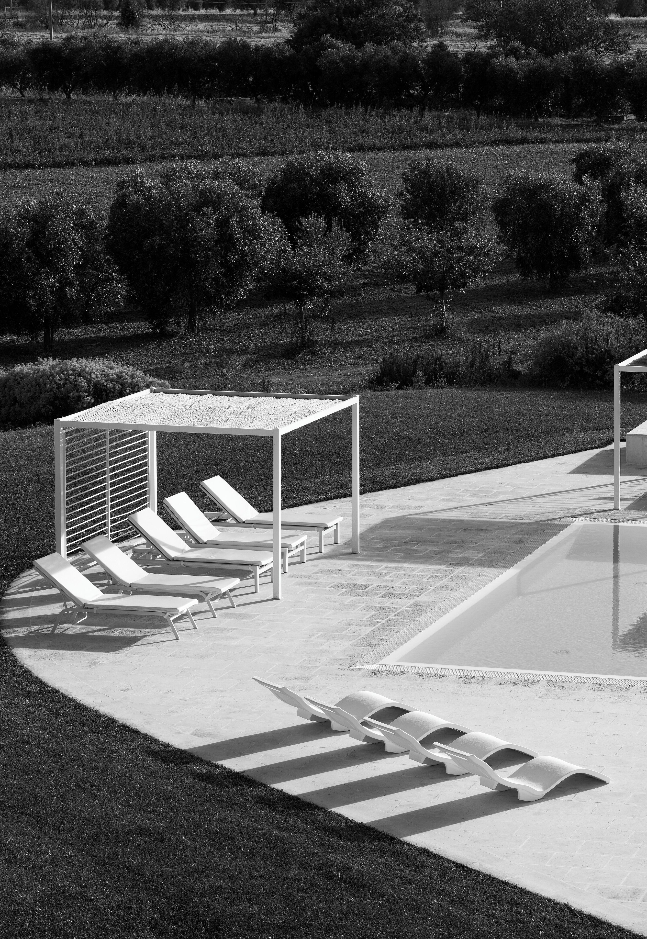 A black and white photo of a poolside area with lounge chairs and a pergola.