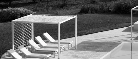 A black and white photo of a poolside area with lounge chairs and a pergola.