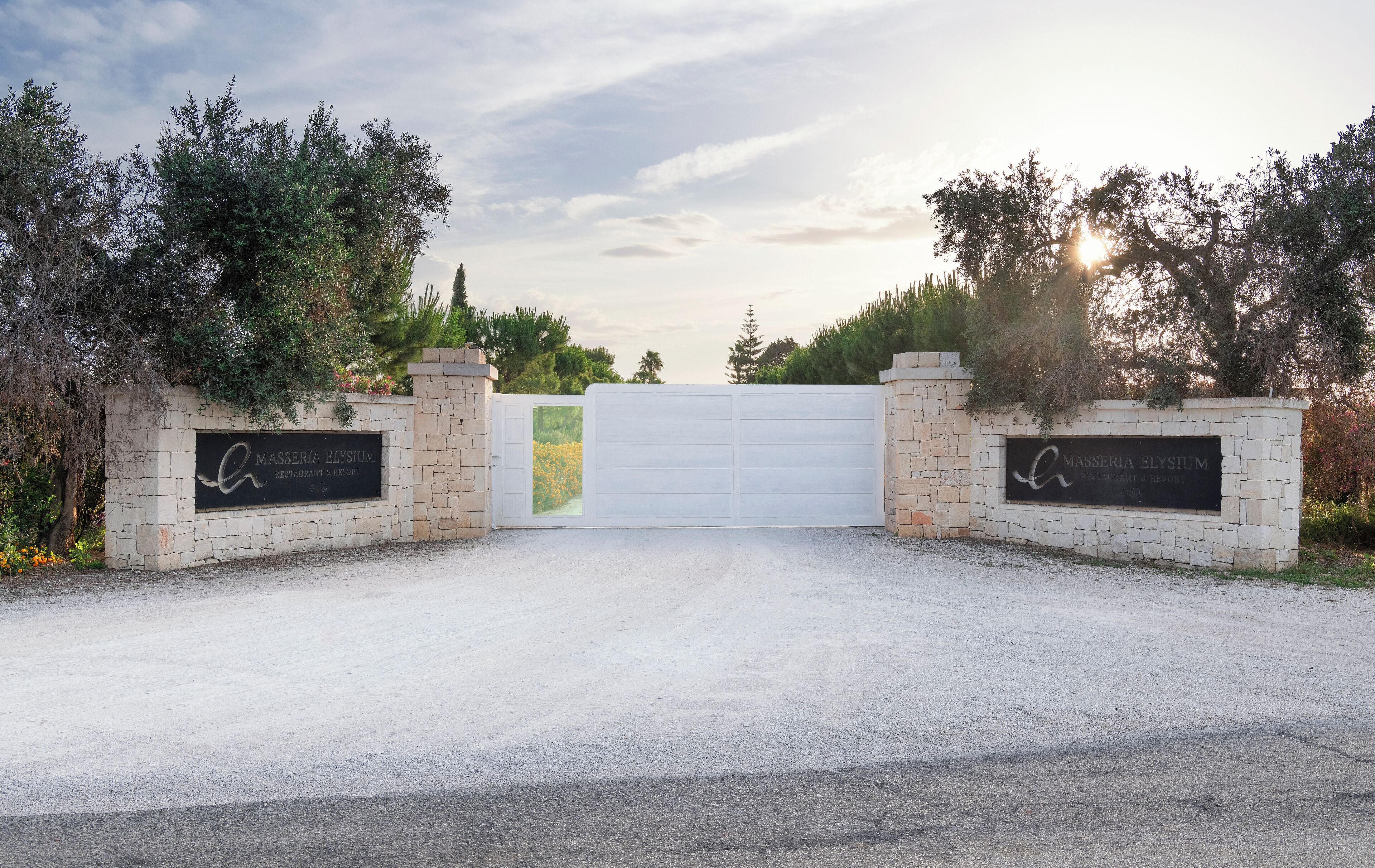 A gated entrance to MASSERIA ELYSIUM restaurant & resort with stone walls and a white gate.
