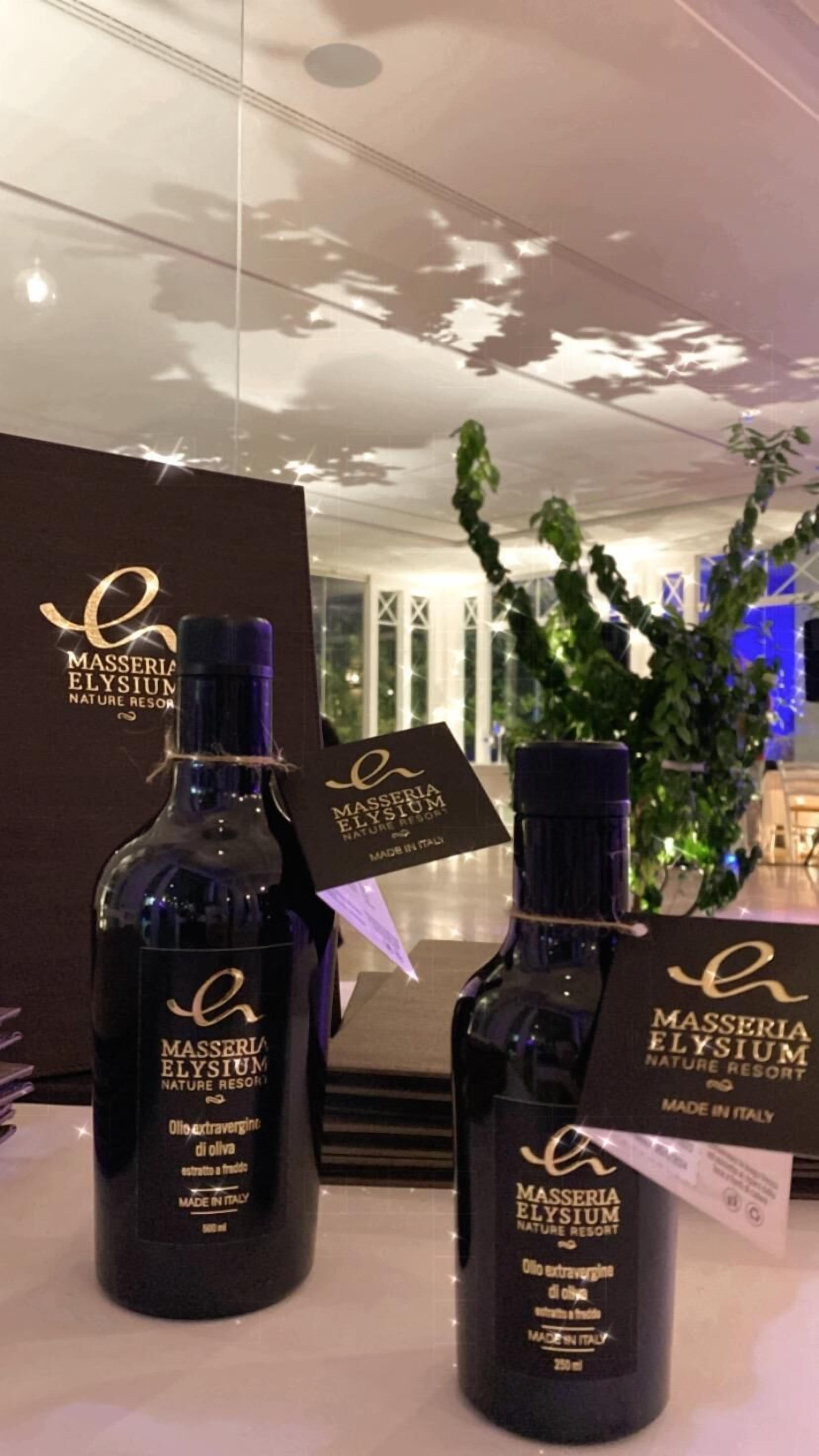 Deux bouteilles d’huile d’olive du MASSERIA ELYSIUM Nature Resort, avec leurs étiquettes et leurs étiquettes d’identification, sont posées sur une table. En arrière-plan, on aperçoit un mur décoré sur le thème de la nature et une plante en pot.