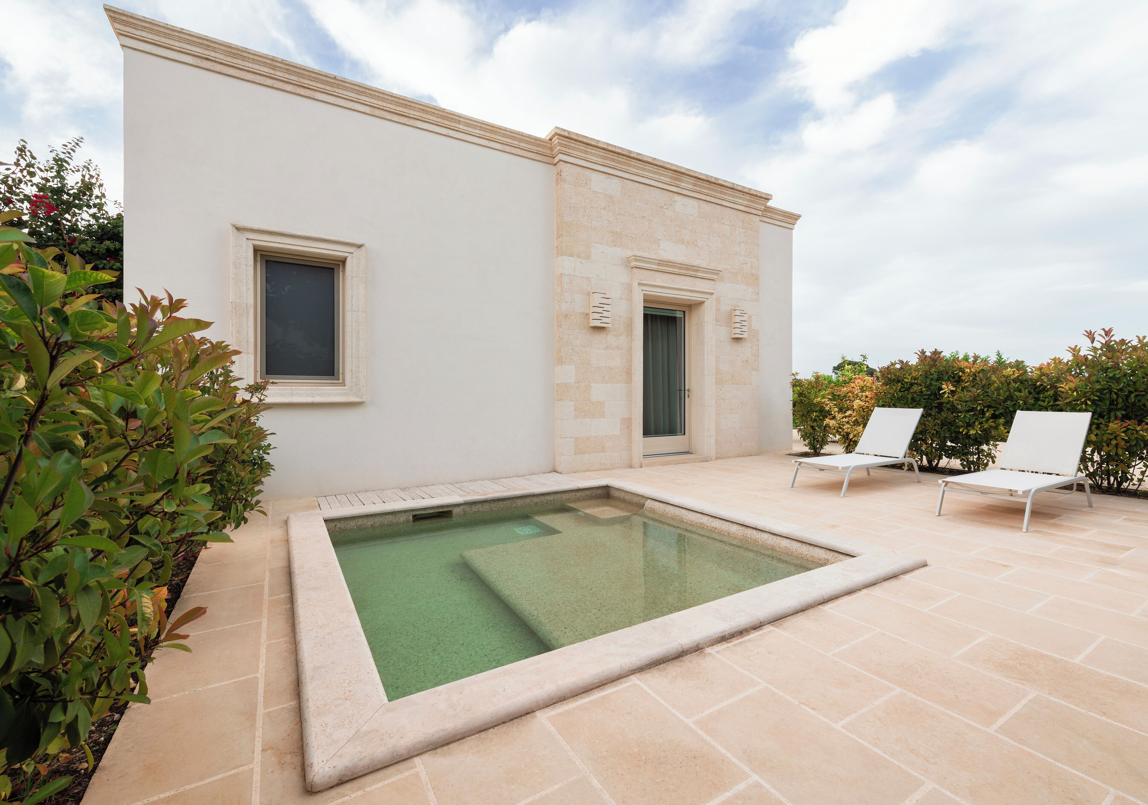 Une piscine avec une terrasse carrelée, deux chaises longues et un bâtiment doté d’une porte et d’une fenêtre.