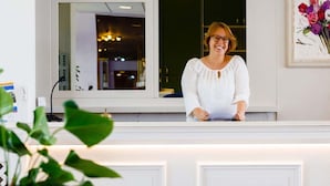 A reception desk with a person standing behind it, a framed picture of flowers on the wall, and a window reflecting interior lighting. - Hotel Royal (Cattolica)