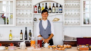 A bar area with shelves of wine bottles and glasses, a bar counter with a pitcher of orange juice, a teapot, and pastries. - Hotel Royal (Cattolica)