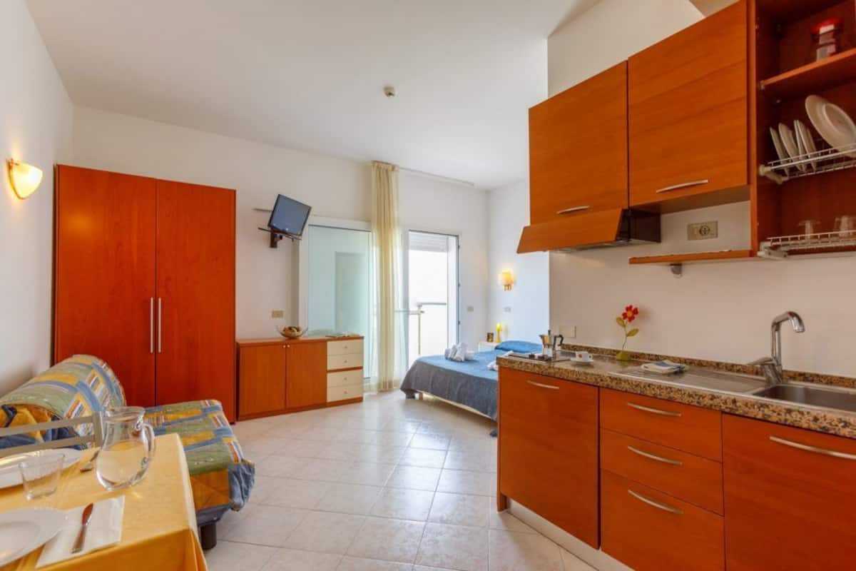 A compact hotel room with a bed, a small table with a pitcher and glasses, a kitchenette with a sink and wooden cabinets, and a TV mounted on the wall.