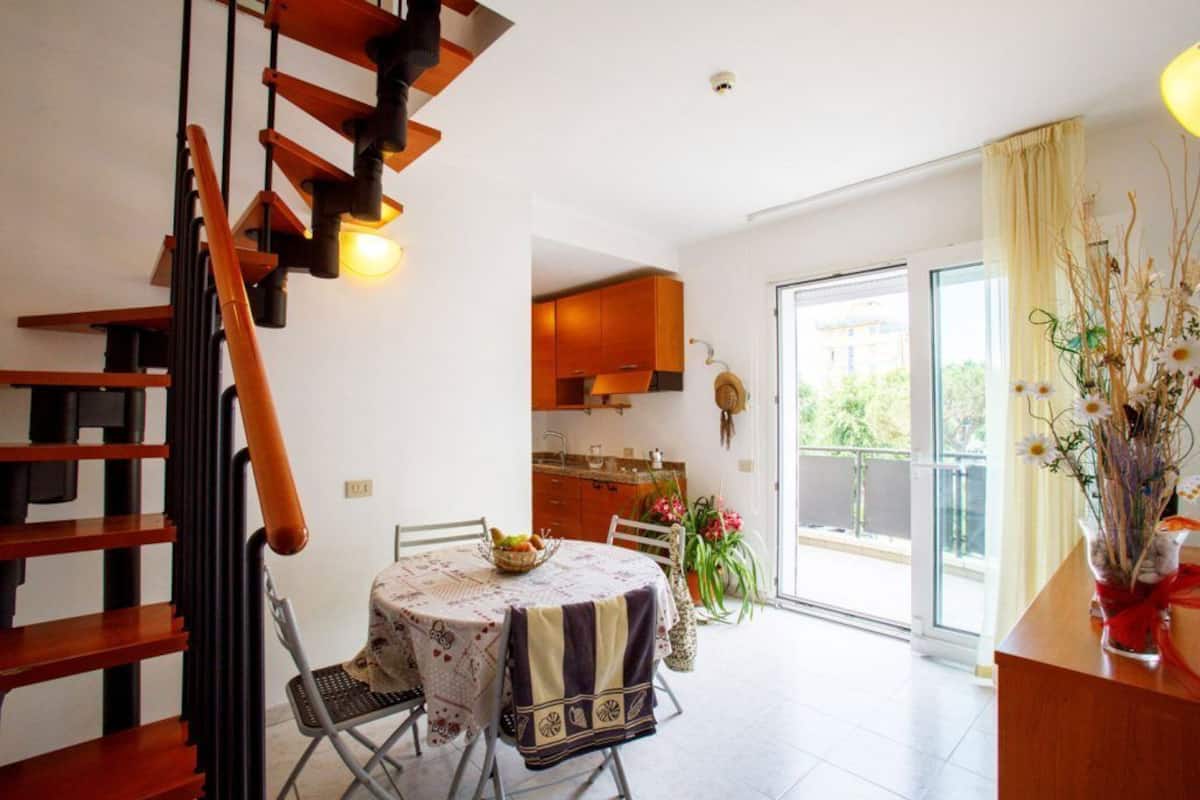 A dining area with a table set for two, a staircase, and a view of the kitchen and balcony.