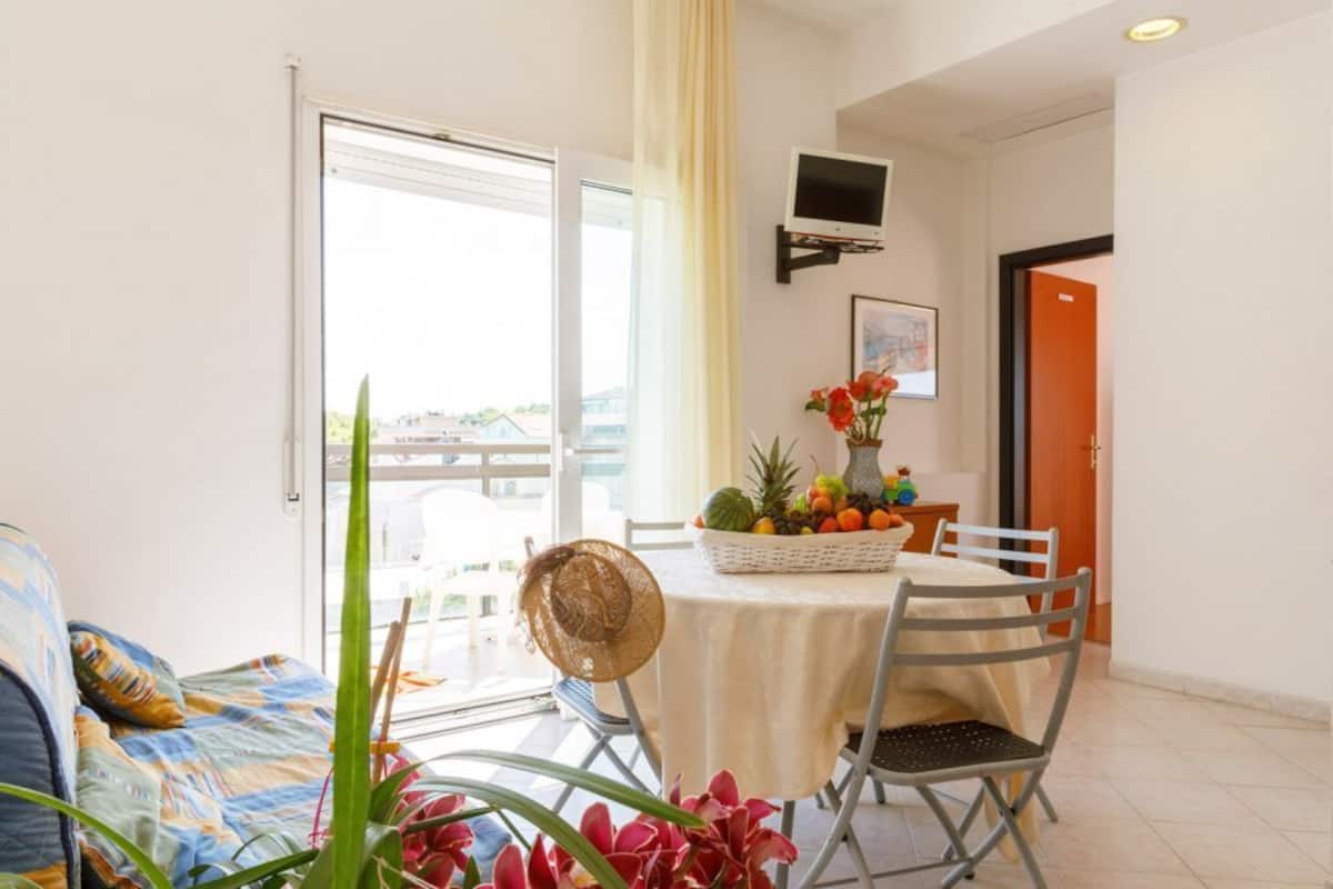 A dining area with a table set for two, a television mounted on the wall, and a balcony door.
