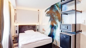 A modern hotel room with a bed, a small desk, a refrigerator, and a large abstract painting on the wall. - Hotel Stadt Grevenbroich (Grevenbroich)