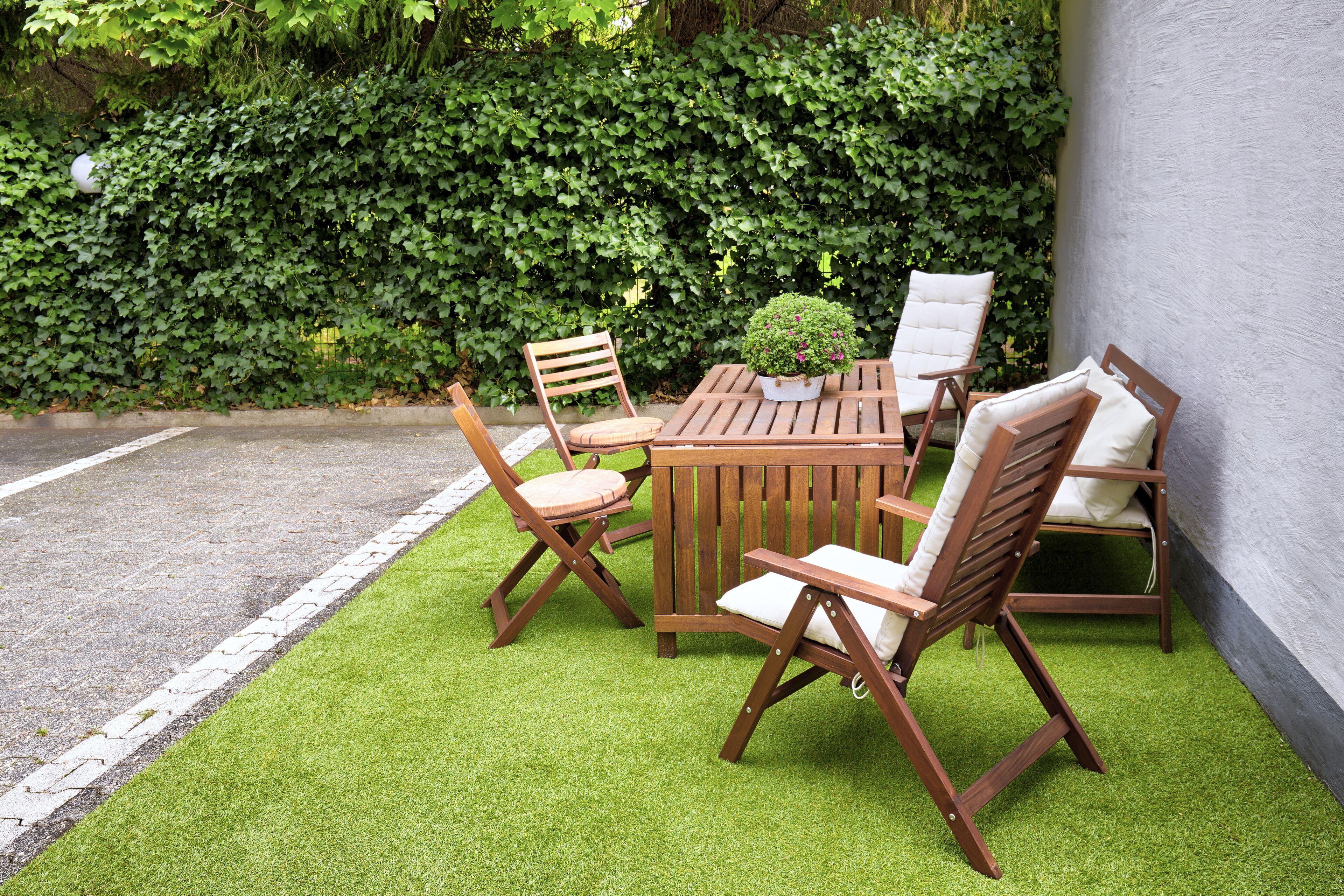A wooden outdoor seating area with a table and chairs, a small potted plant, and a hedge in the background.