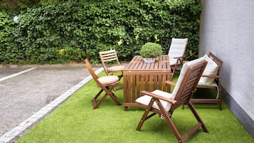 Un área de estar al aire libre con madera, que incluye una mesa, sillas, una planta pequeña en maceta y un seto al fondo.