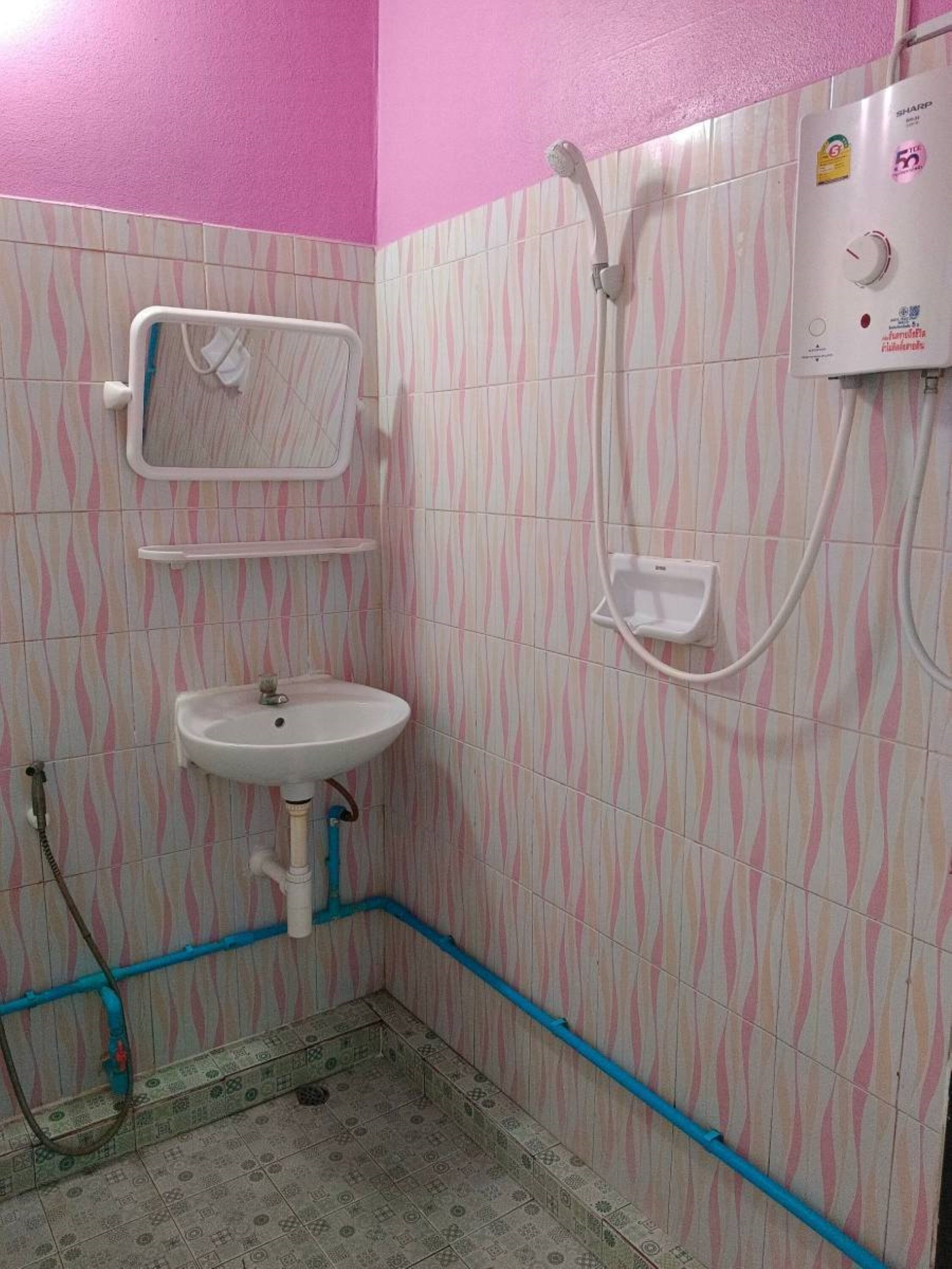 Casa de banho com parede de azulejos listrada em rosa e branco, uma pia branca, um chuveiro manual e um termoacumulador.
