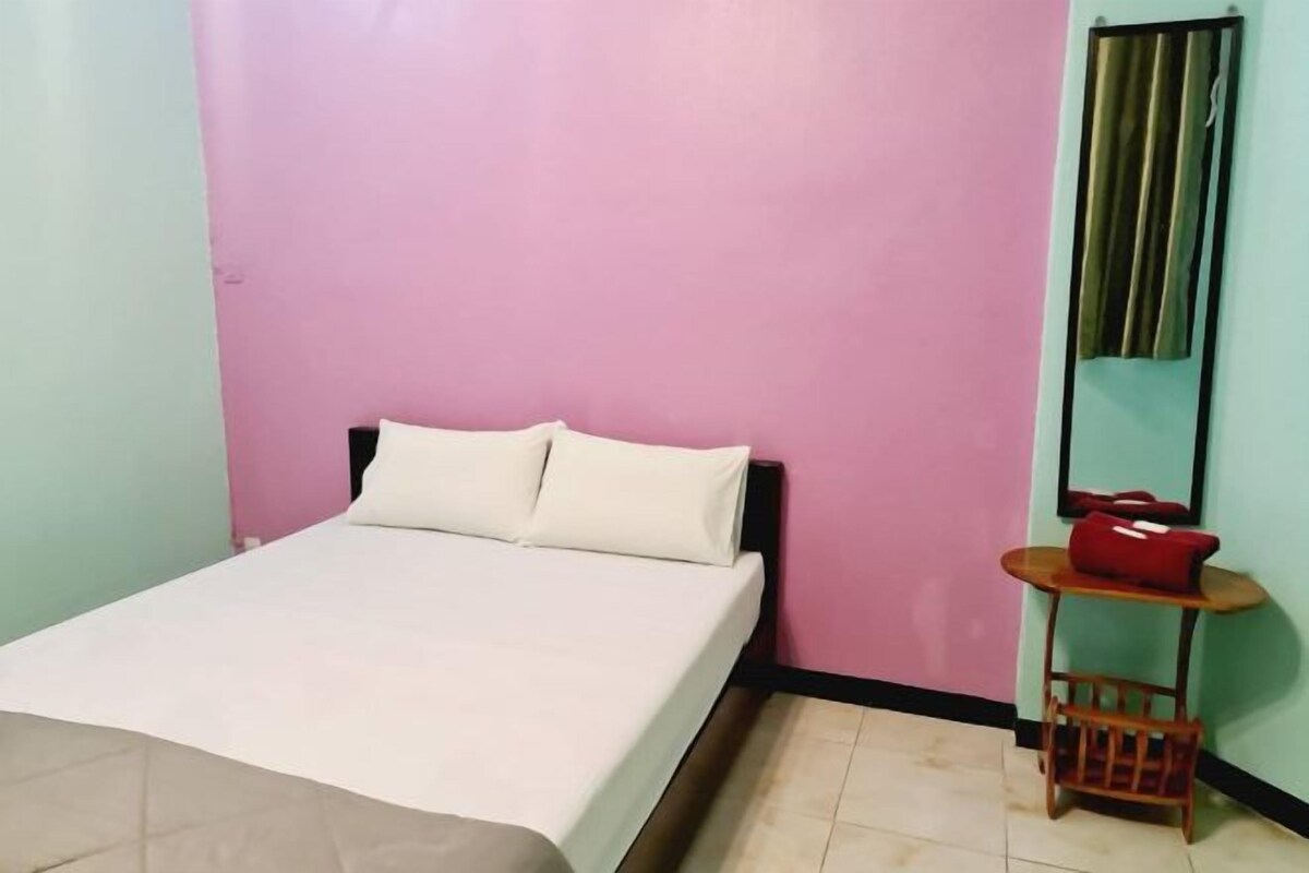 A single bed with a white mattress and pillows, a small wooden table with a red bag, and a mirror on the wall.