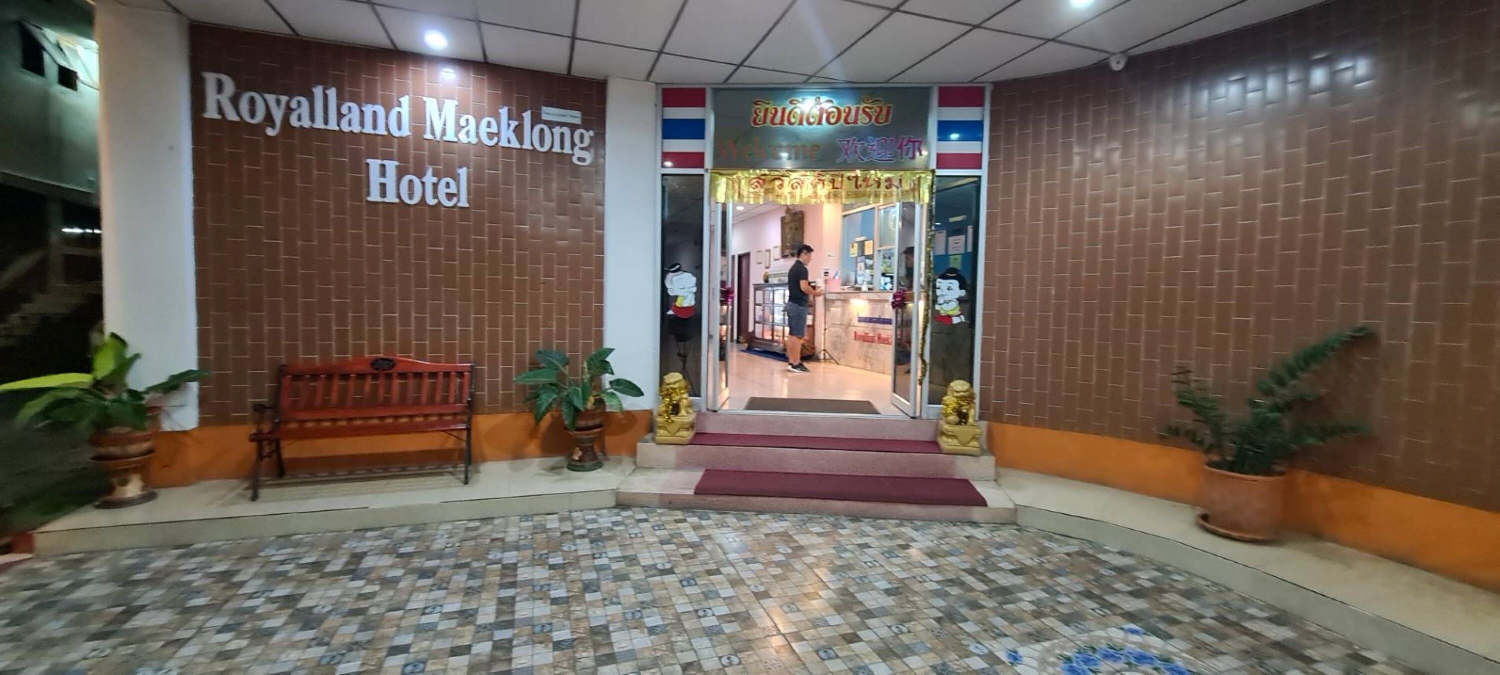 The entrance to the Royalland Maeklong Hotel with a tiled floor and potted plants.